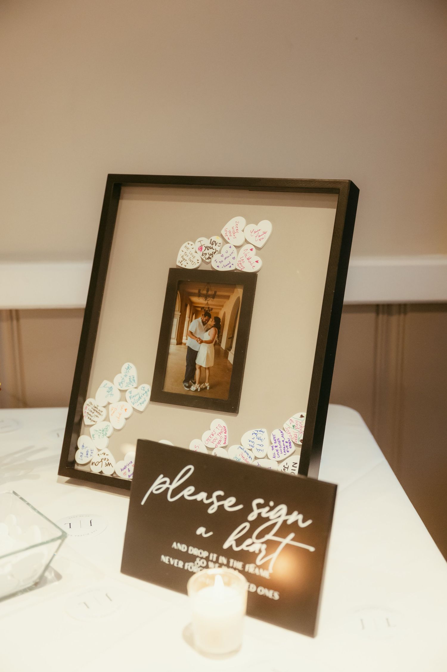 Wedding guest book frame with heart-shaped sign-in cards surrounding a couple photo, next to a please sign a heart sign.