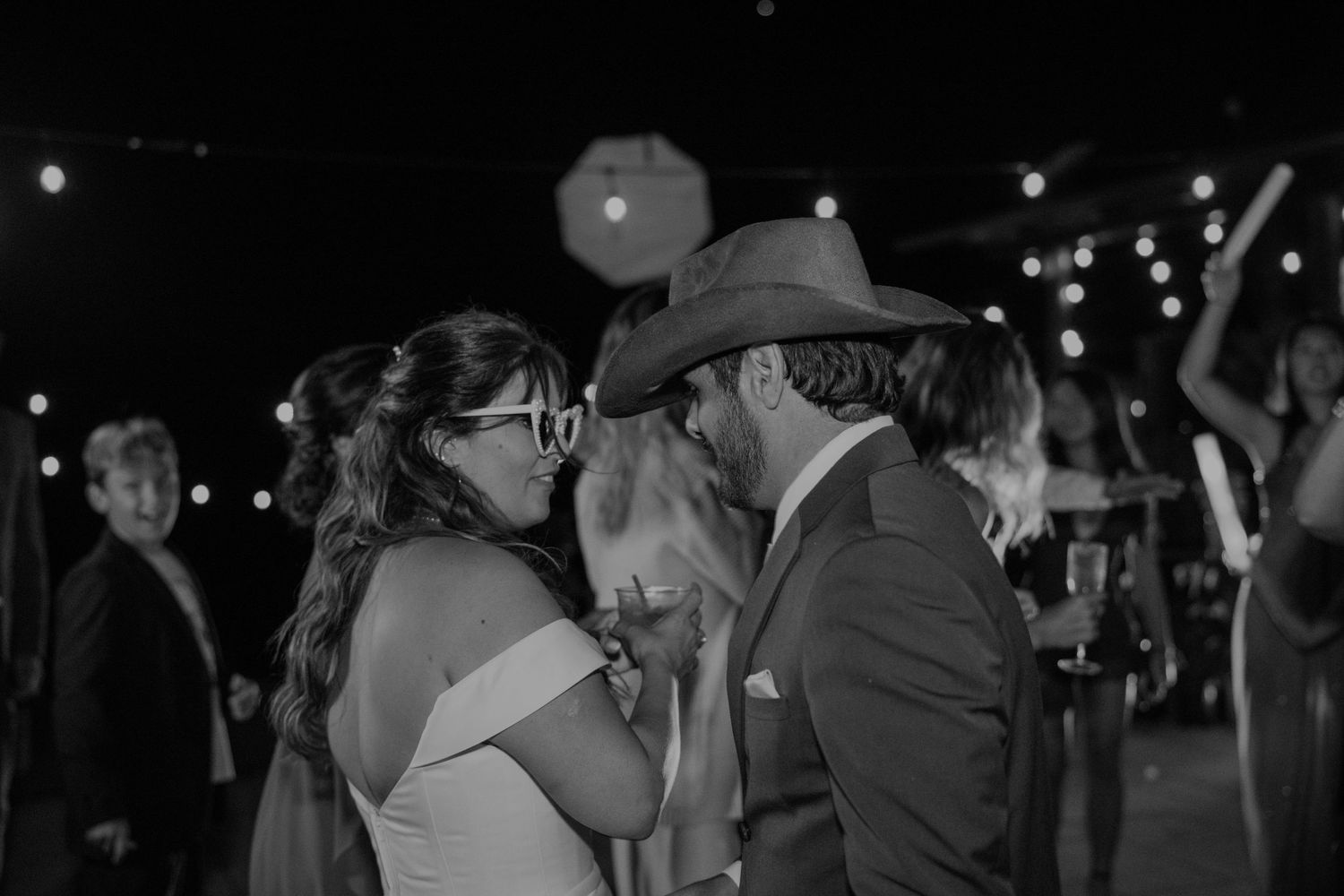 Black and white photo of a couple sharing a romantic moment on a wedding dance floor with string lights.
