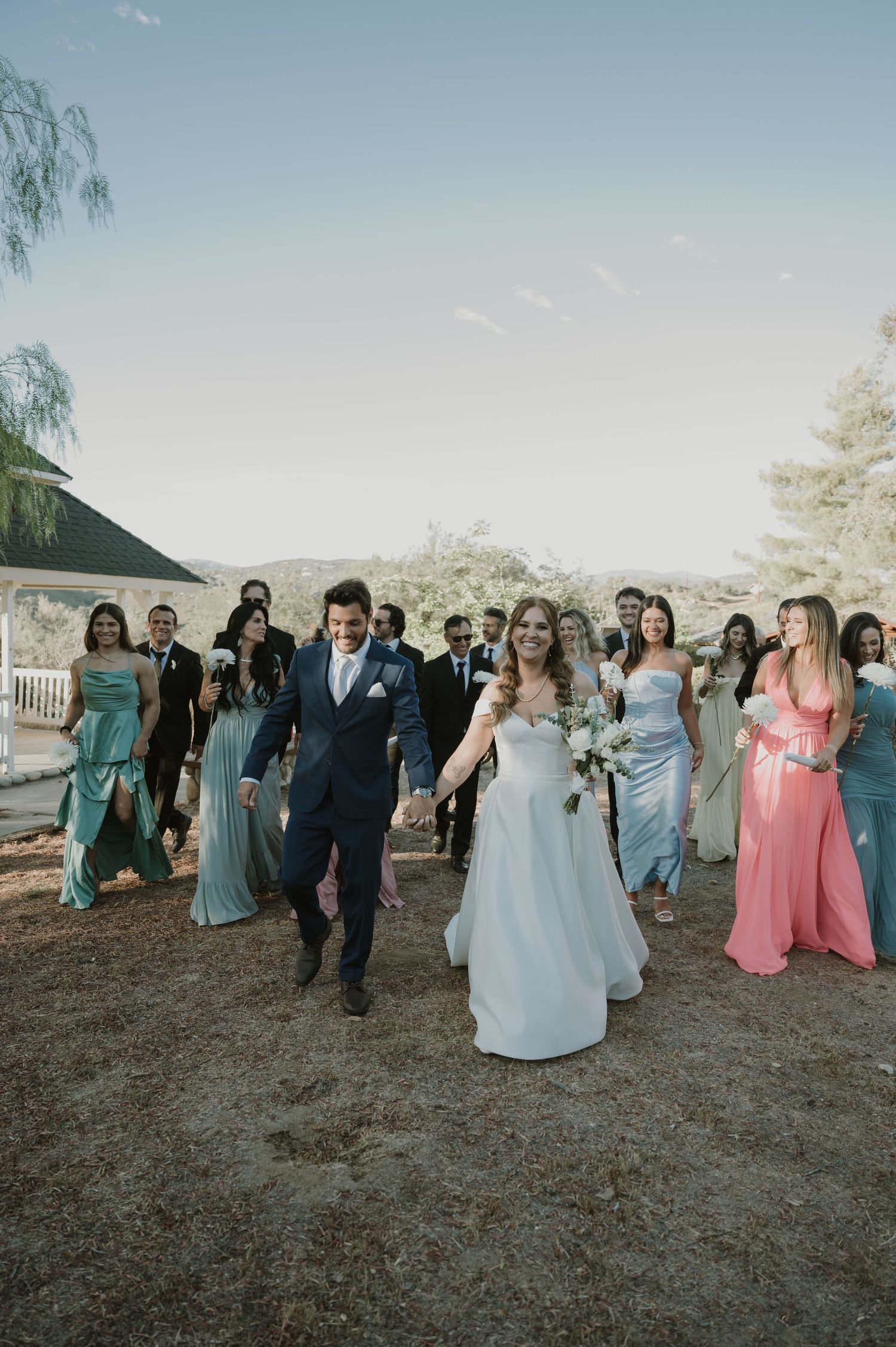 A bride and groom lead their wedding party across an outdoor venue, bridesmaids in pastel gowns.