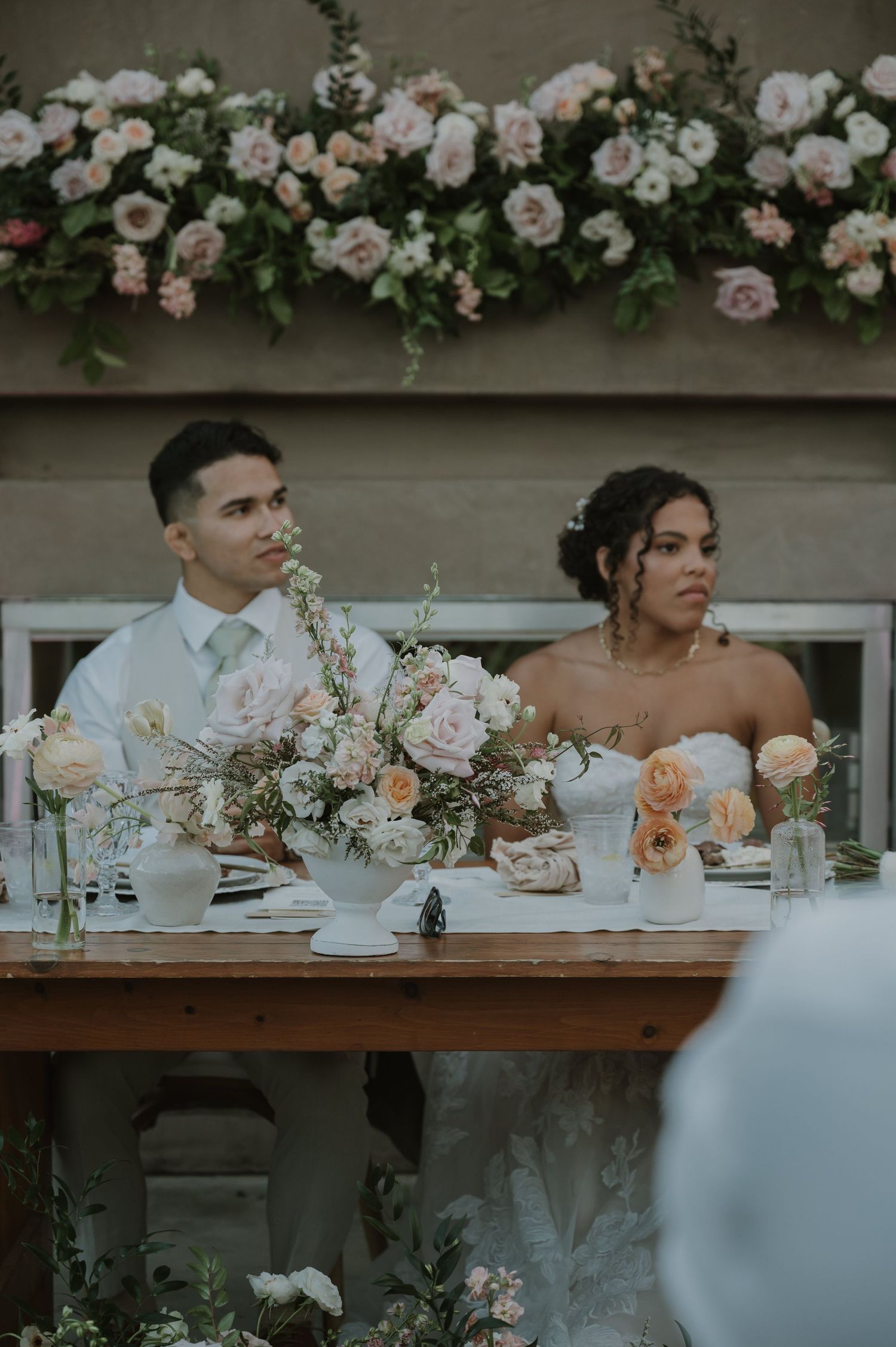 Bride and groom seated at a floral-adorned sweetheart table with soft pink roses and candlelight at their wedding reception.