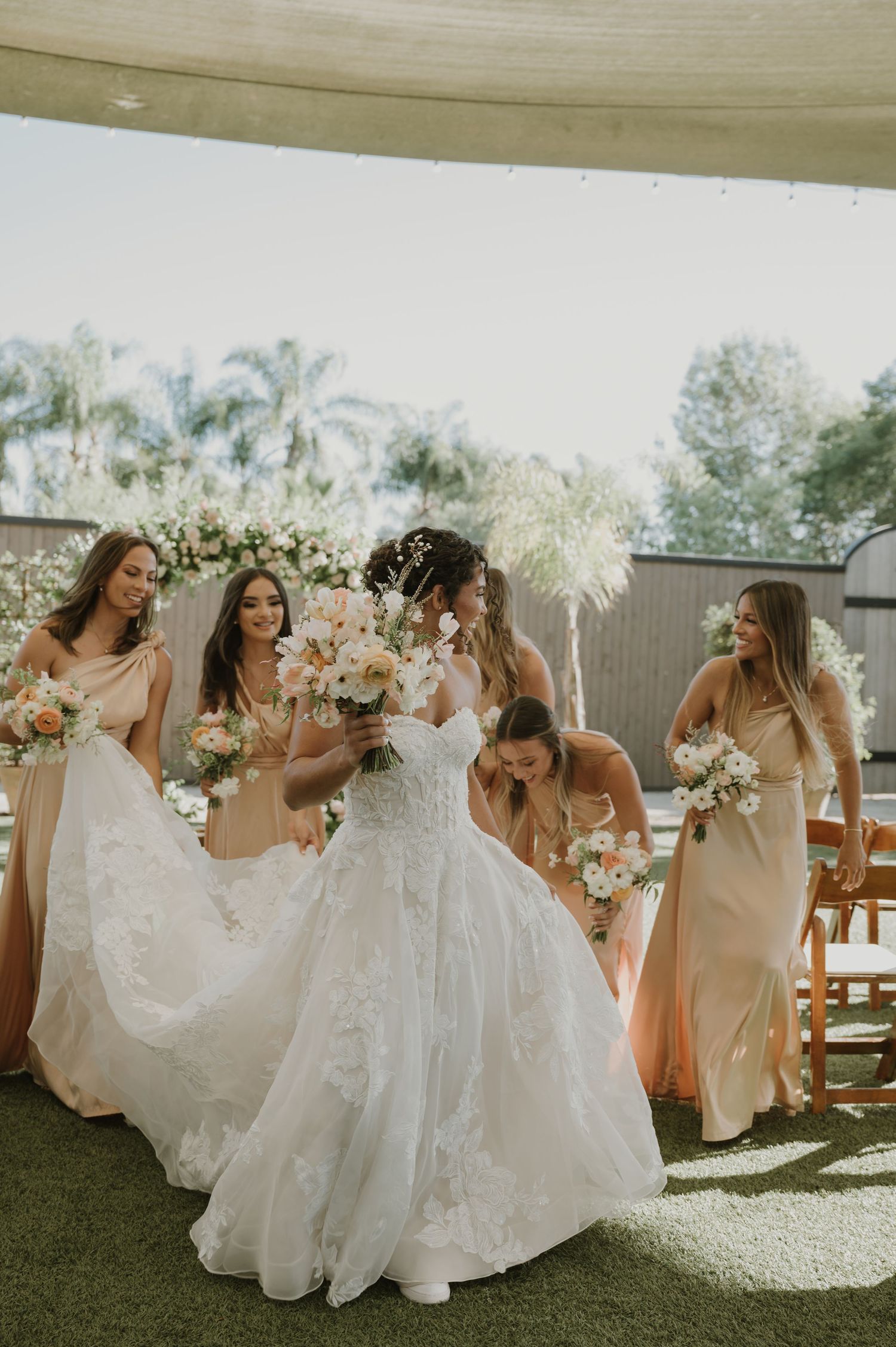Bride in lace ballgown holds floral bouquet surrounded by bridesmaids in champagne dresses at outdoor wedding ceremony.