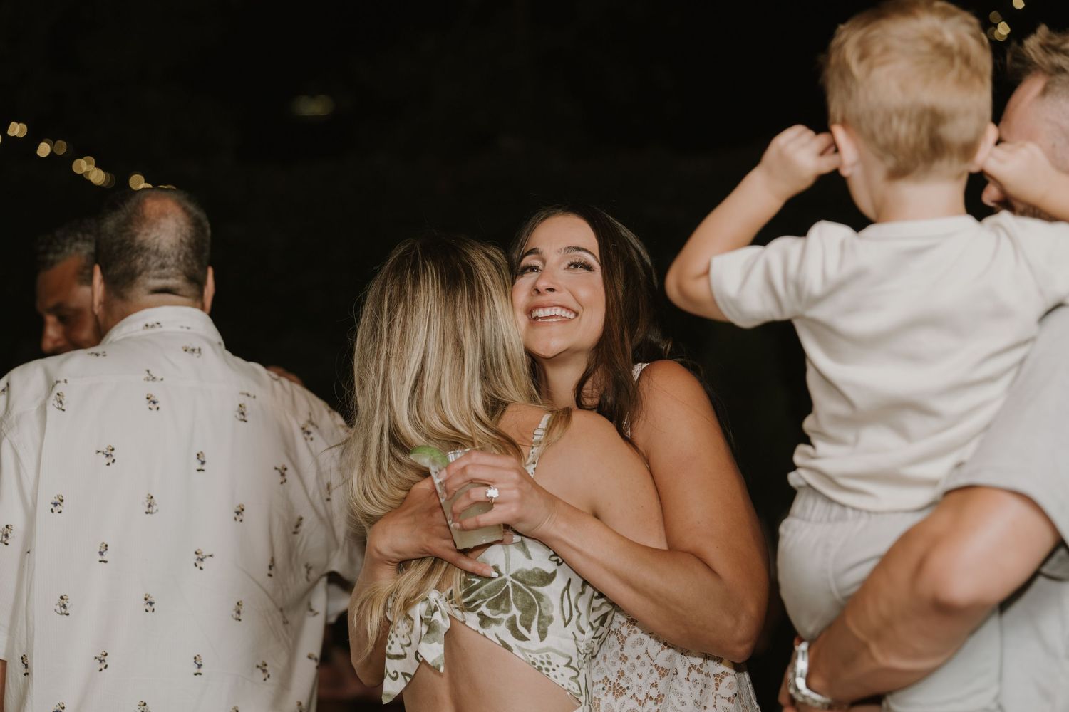 Two women share a warm hug at an outdoor wedding reception while a child is held nearby.