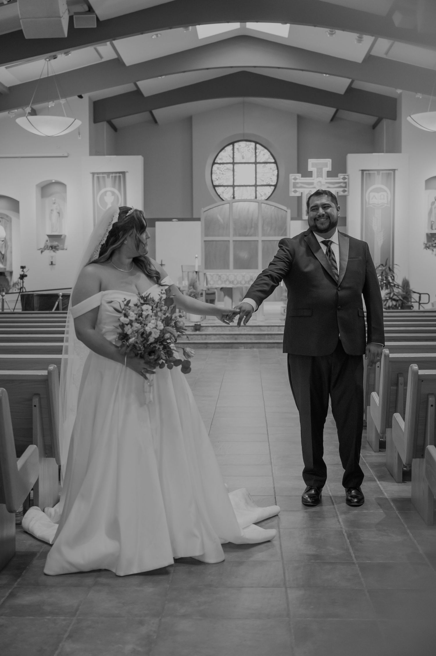 Black and white photo of a bride and groom holding hands in a church aisle during their wedding ceremony.