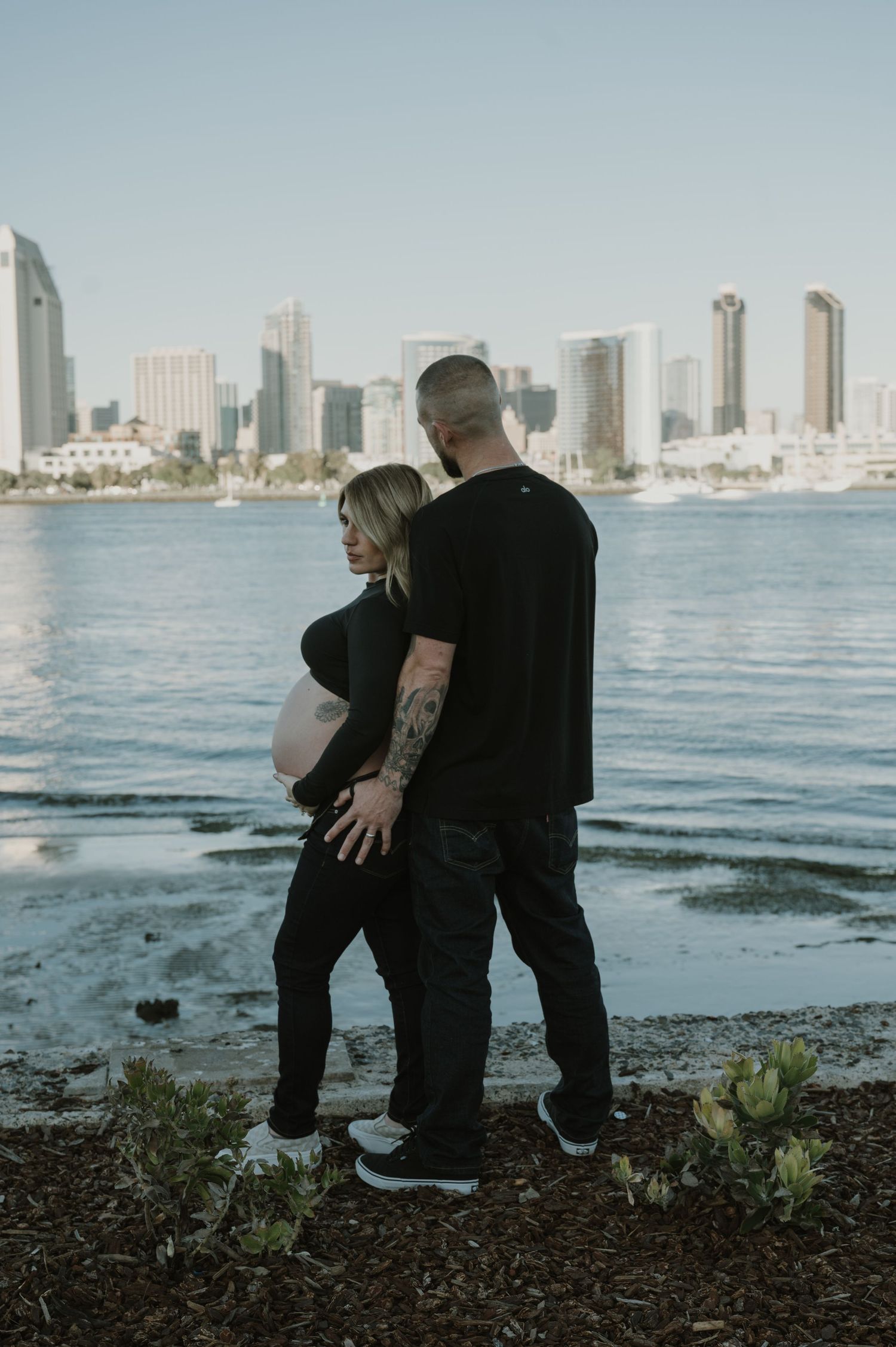 Couple sitting by waterfront with urban skyline visible across the water.