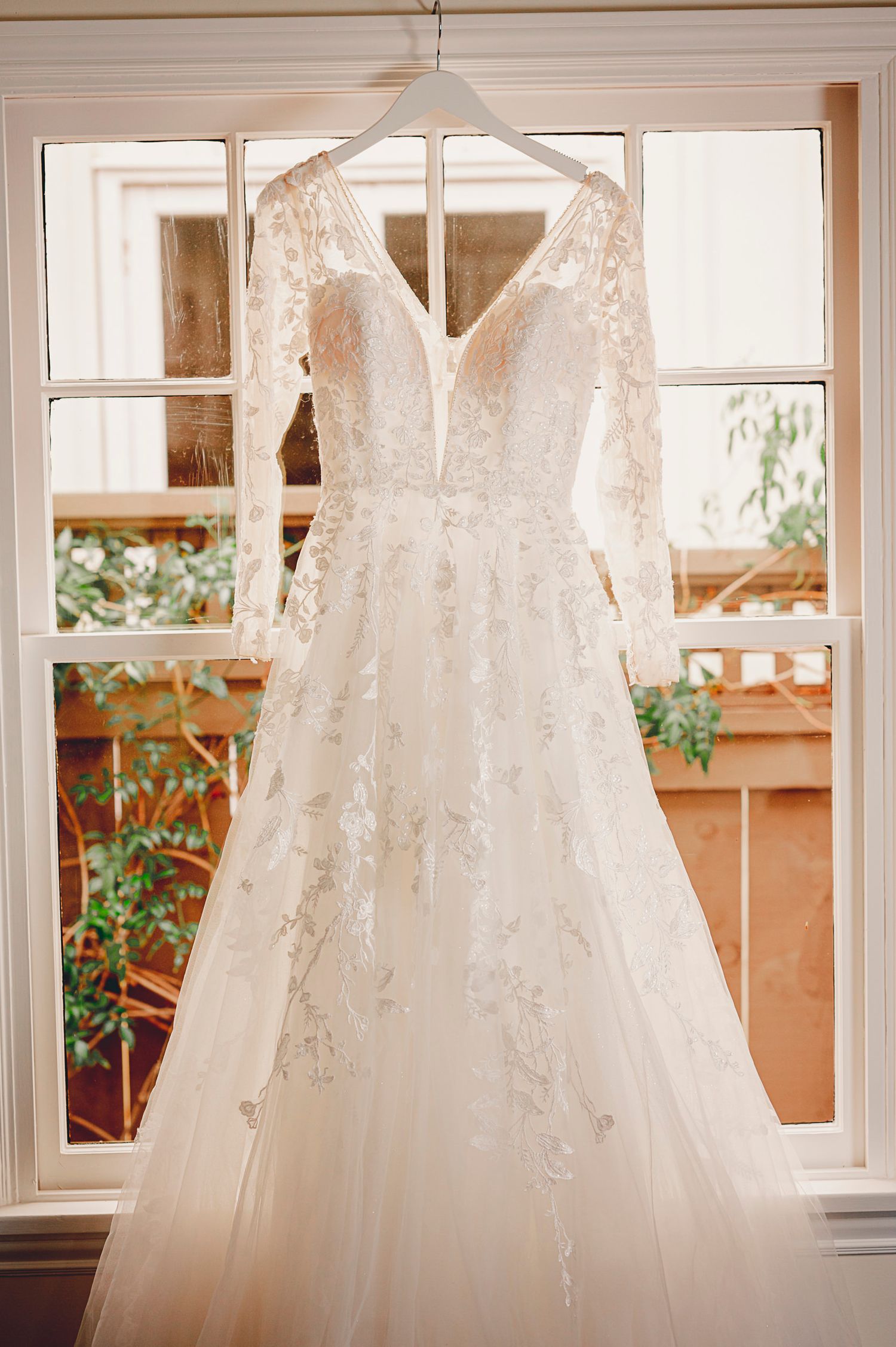 White lace wedding dress with plunging neckline hanging in window with natural light.