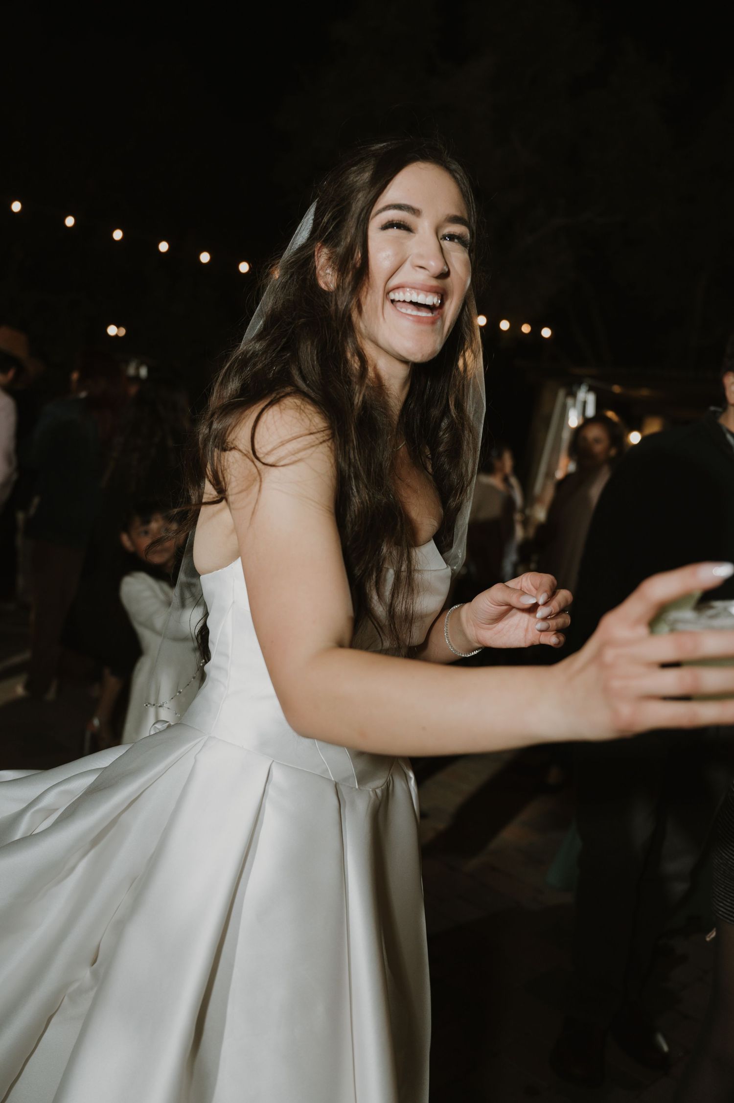 Bride in a white wedding dress laughs and dances joyfully at a nighttime reception.
