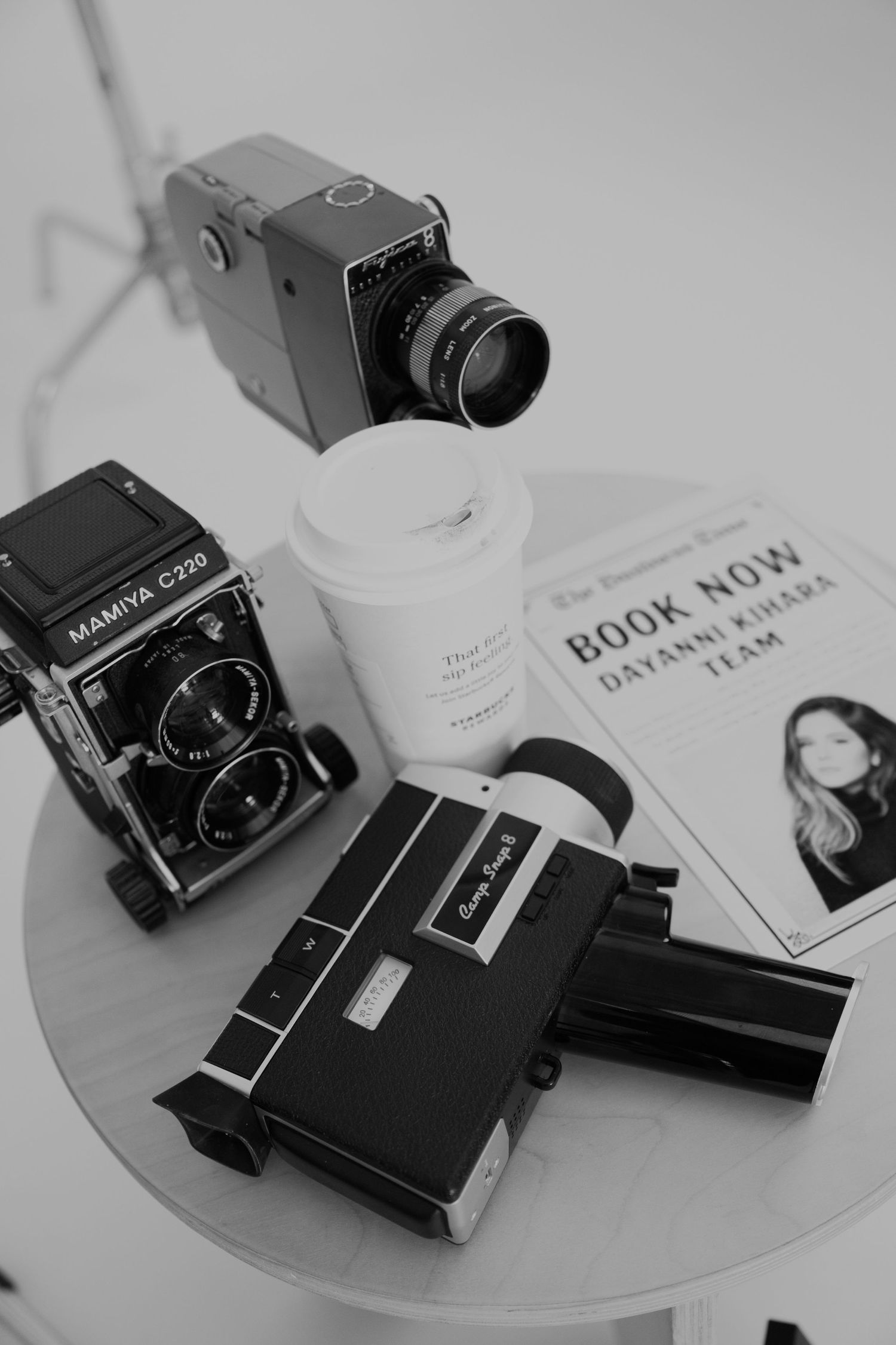 A vintage Mamiya medium format film camera displayed alongside books and papers on a white surface in monochrome.