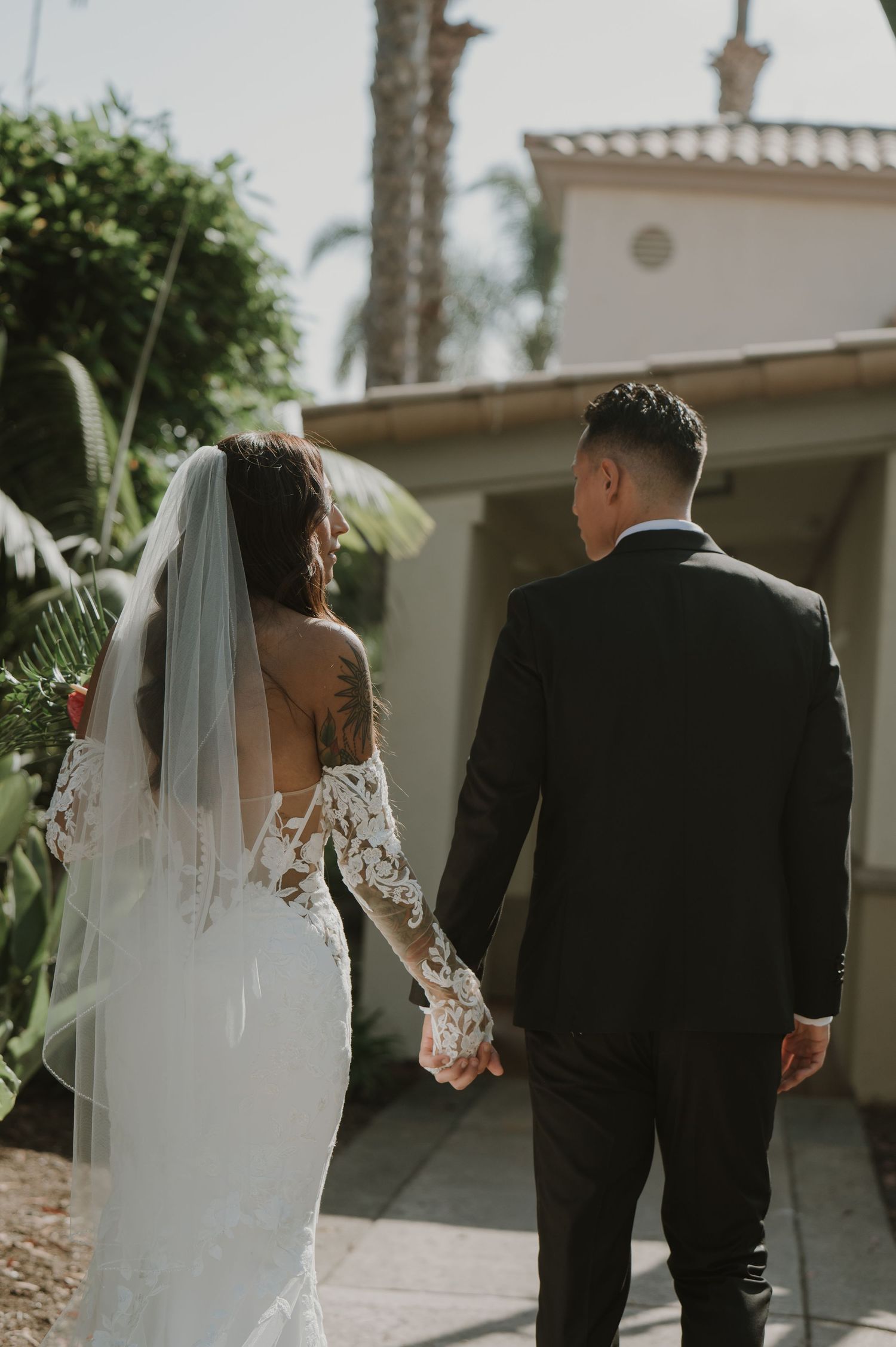 Wedding couple walking together along a path with palm trees and white buildings.