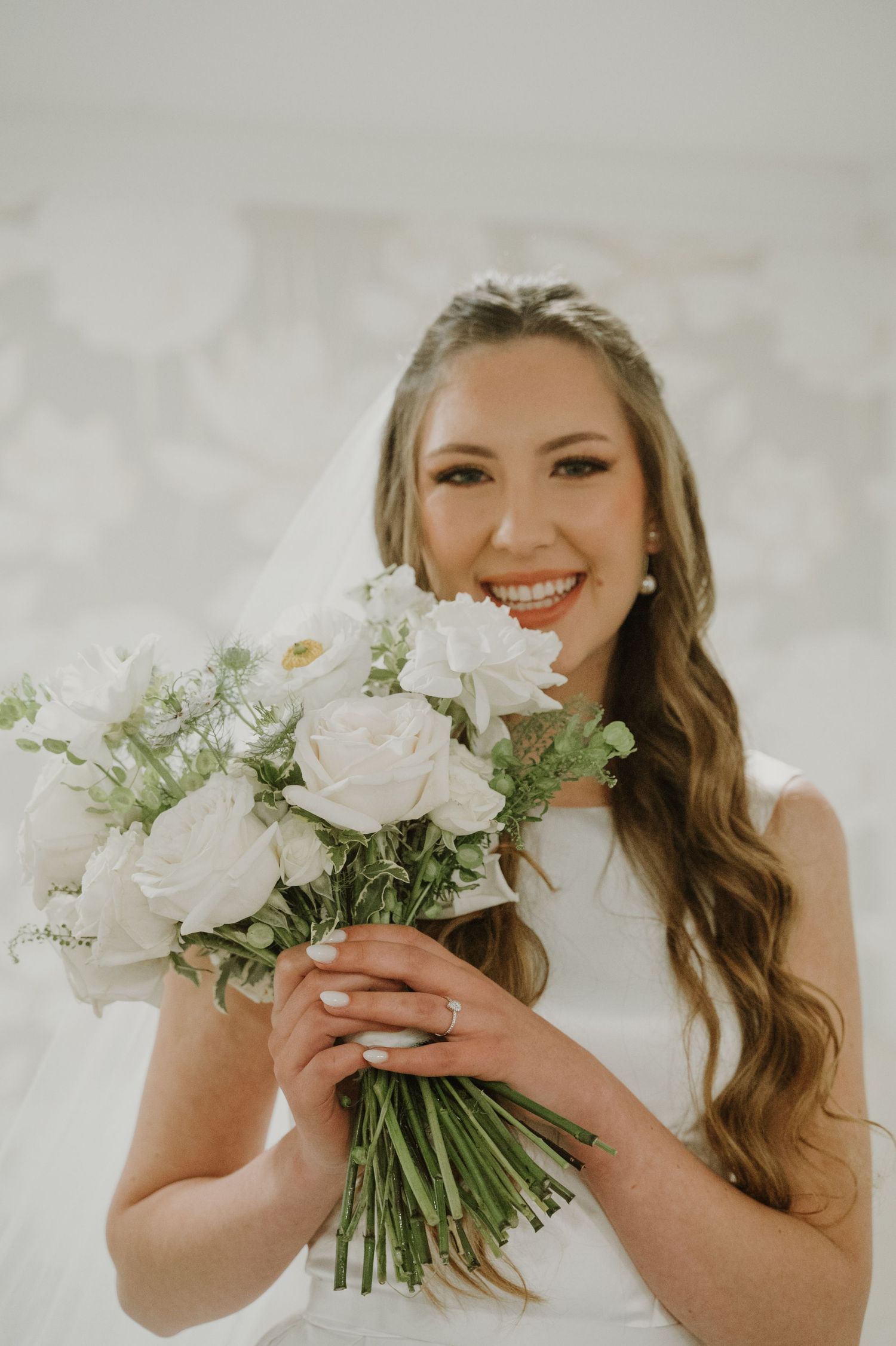 Smiling bride in a white gown holding a bouquet of white roses and greenery.