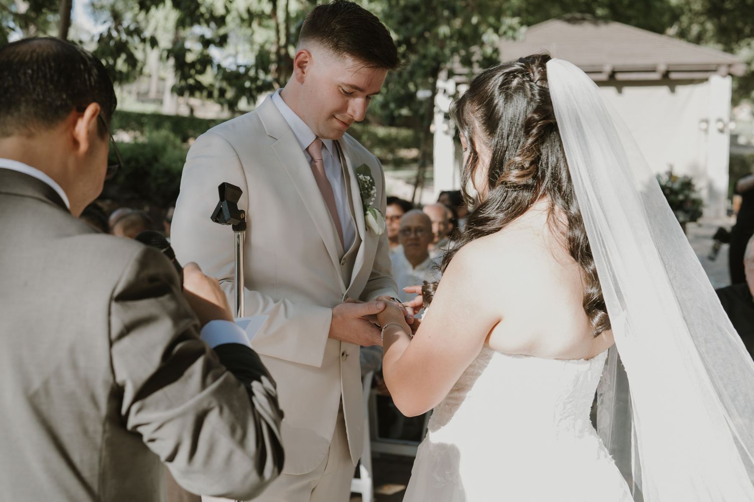Groom in beige suit places ring on brides finger during outdoor wedding ceremony exchange.