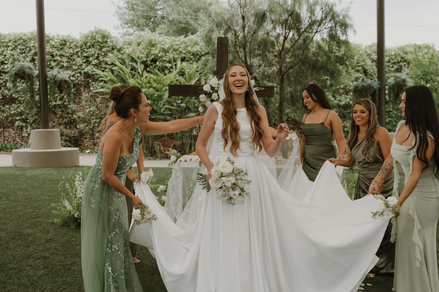 Bride in white gown laughs with bridesmaids in sage green dresses holding her skirt in an outdoor garden setting.