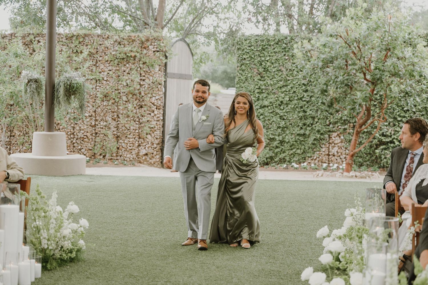 Couple walks down outdoor wedding aisle on green lawn, man in gray suit, woman in sage green gown, floral backdrop.