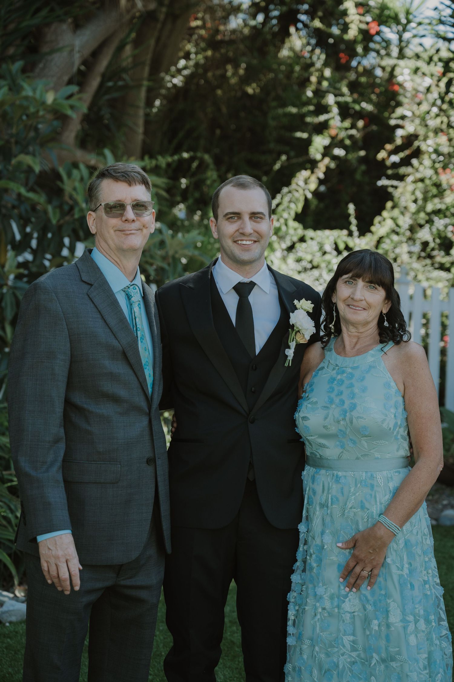 Groom in black suit poses with parents in formal attire at outdoor wedding ceremony.