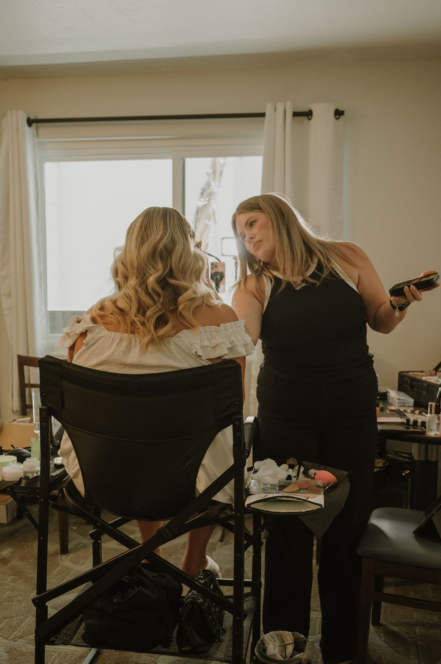 A makeup artist in black applies finishing touches to a blonde bride sitting in a director's chair during wedding prep.