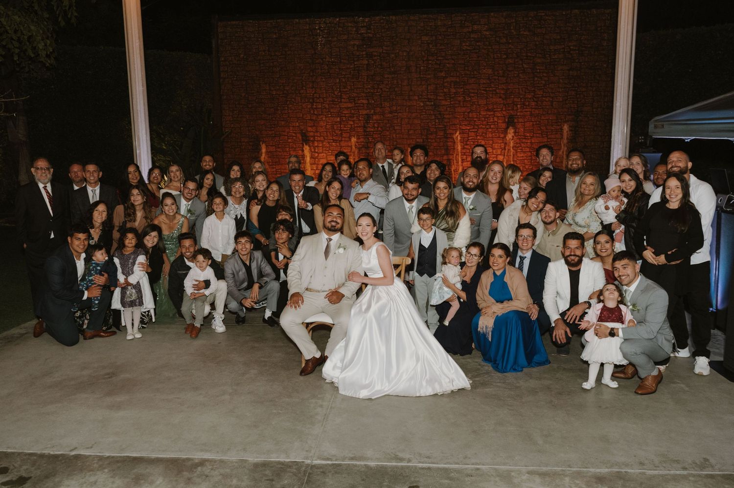 Large wedding group photo with bride and groom seated at center, surrounded by family and guests outdoors at night.