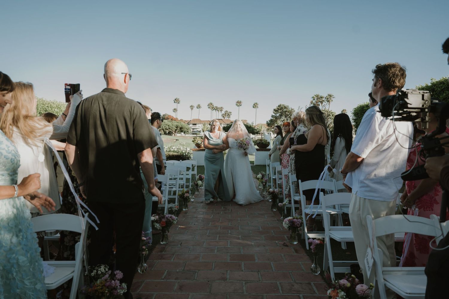 Bride walks down brick aisle with escort at outdoor wedding ceremony, guests seated in white chairs under clear blue sky.