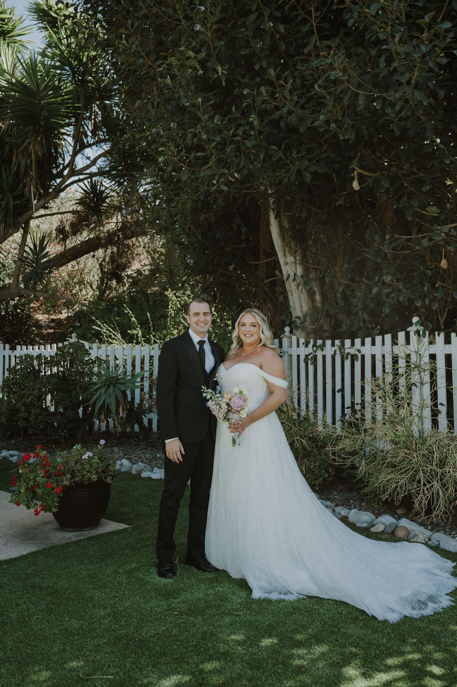 Bride and groom posing together in a garden with white picket fence, bride in off-shoulder gown holding bouquet.