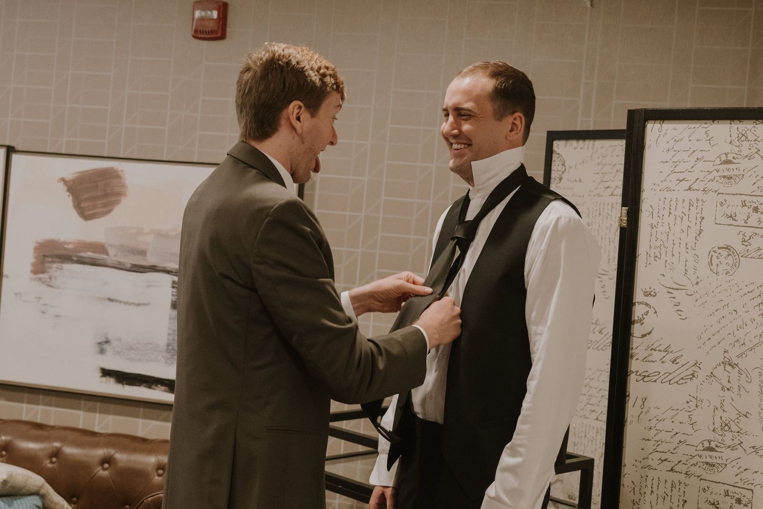 Two men in formal attire help each other with a tie before a wedding ceremony indoors.