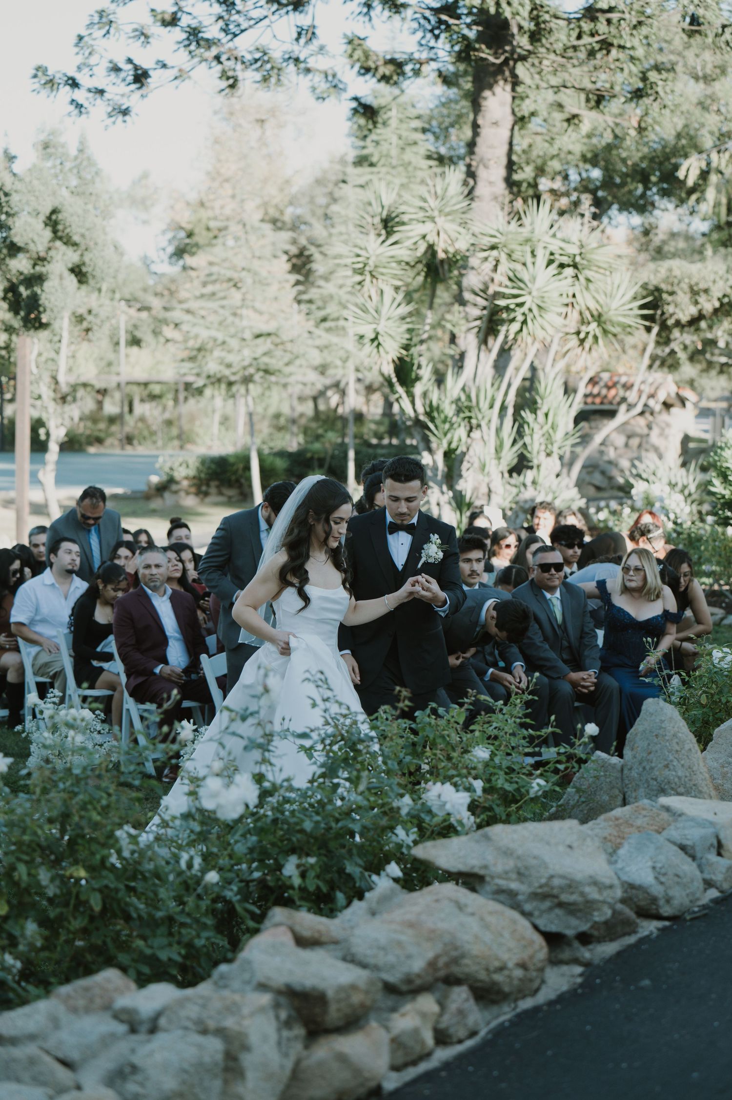 A wedding ceremony takes place in a lush garden setting with stone pathways and gathered guests.