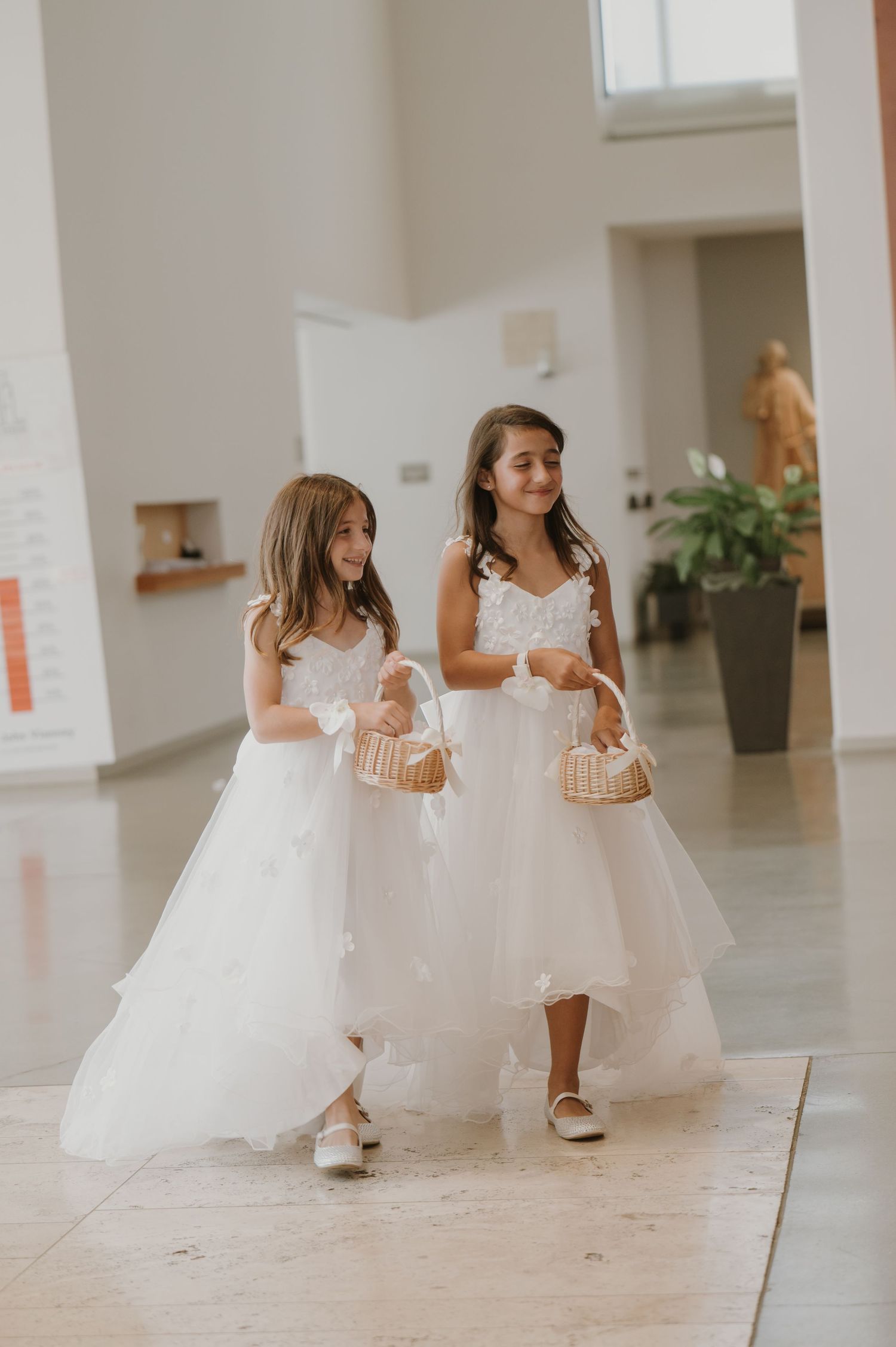 Two flower girls in white tulle dresses carry wicker baskets while walking down a bright church hallway.