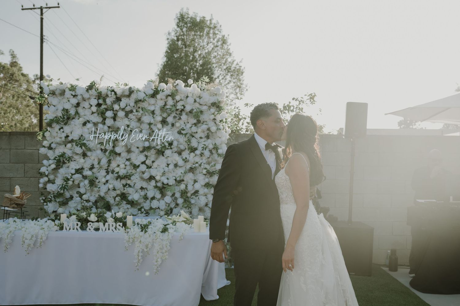 Bride and groom share a kiss in front of a white floral wall backdrop at an outdoor wedding ceremony.