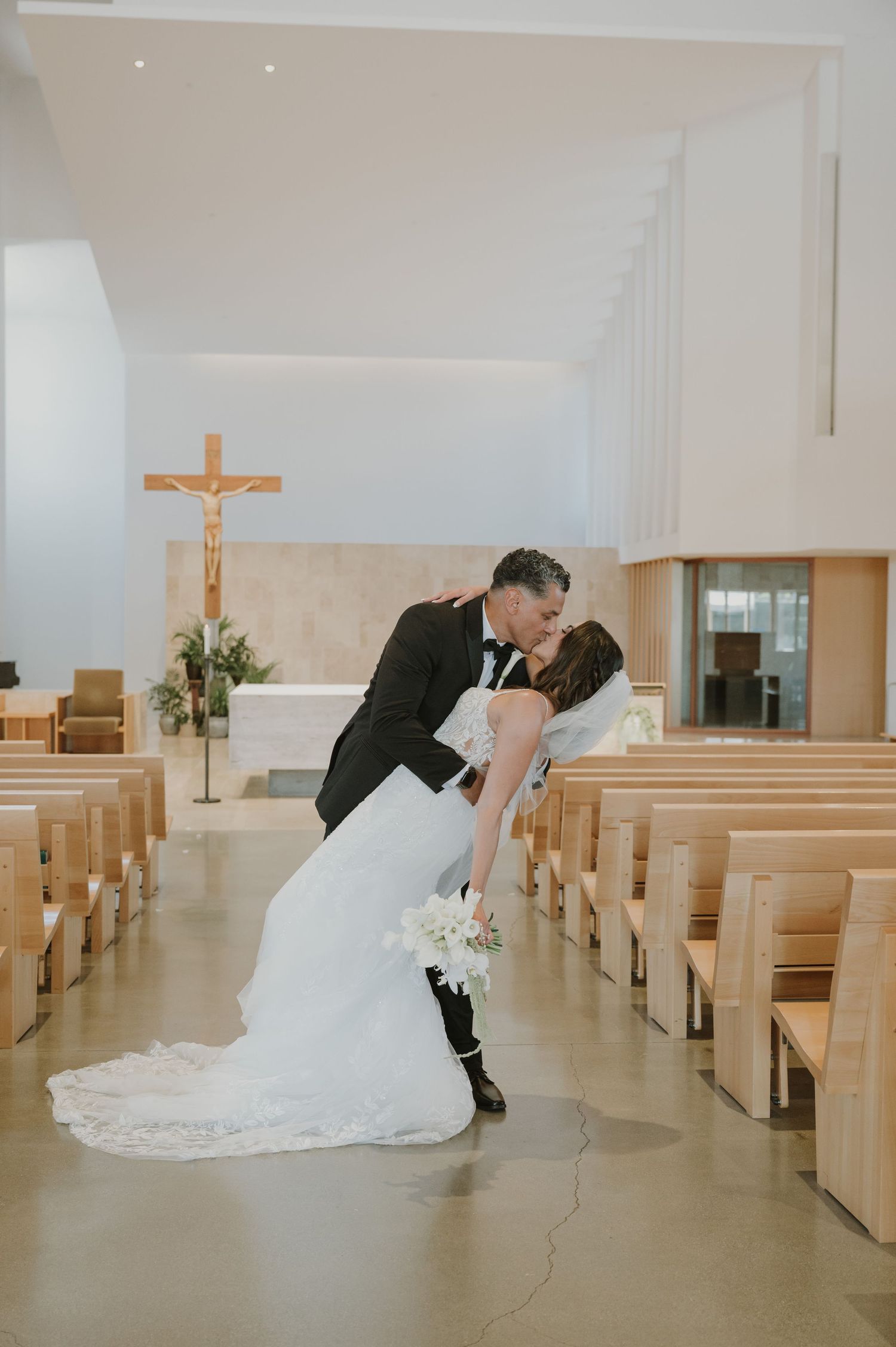 Bride and groom share a romantic dip kiss in a modern Catholic church aisle, with wooden pews and crucifix altar backdrop.