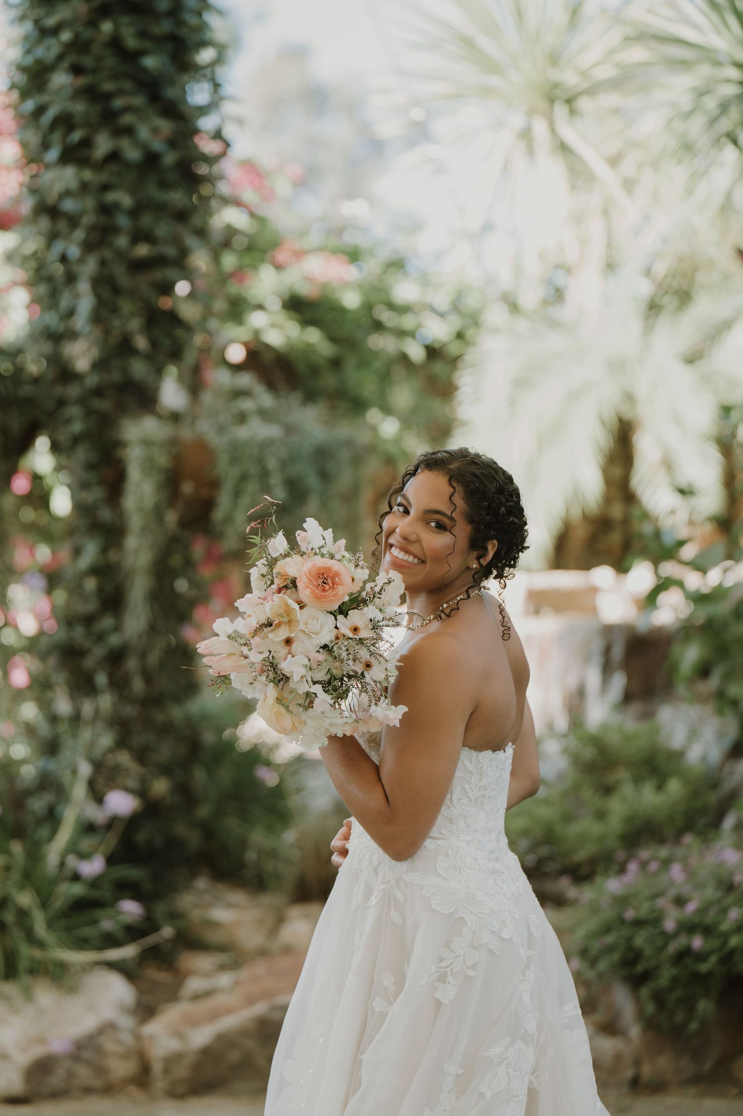 Smiling bride in white strapless gown holding peach floral bouquet in lush garden setting.