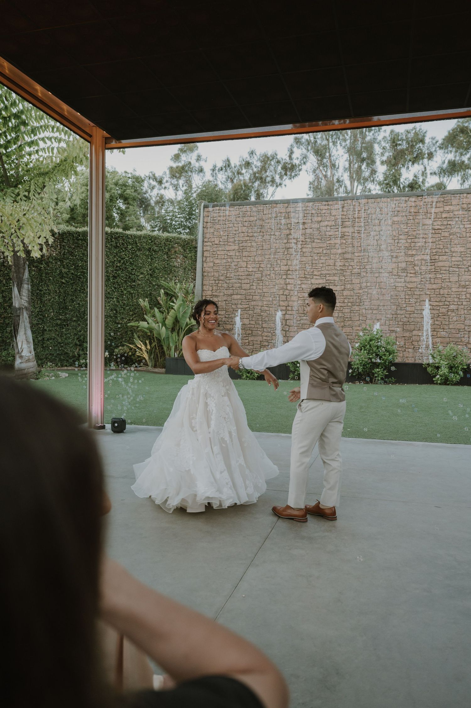 Bride and groom share their first dance outdoors under a covered patio surrounded by lush greenery.
