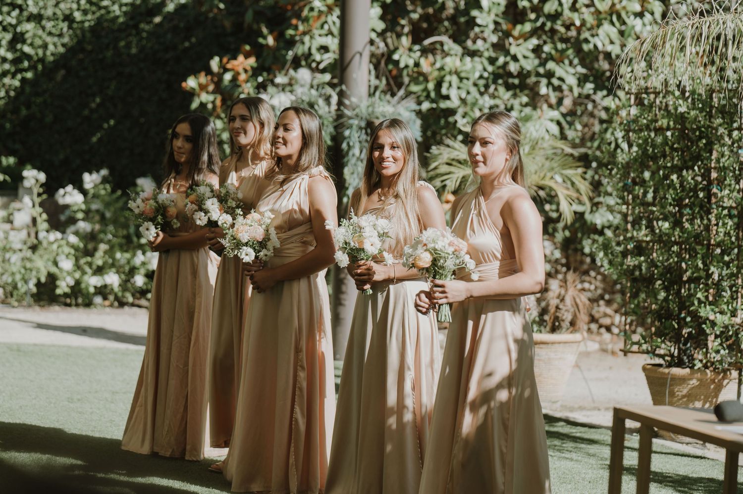 Four bridesmaids in champagne gowns holding white floral bouquets stand outdoors surrounded by lush greenery.