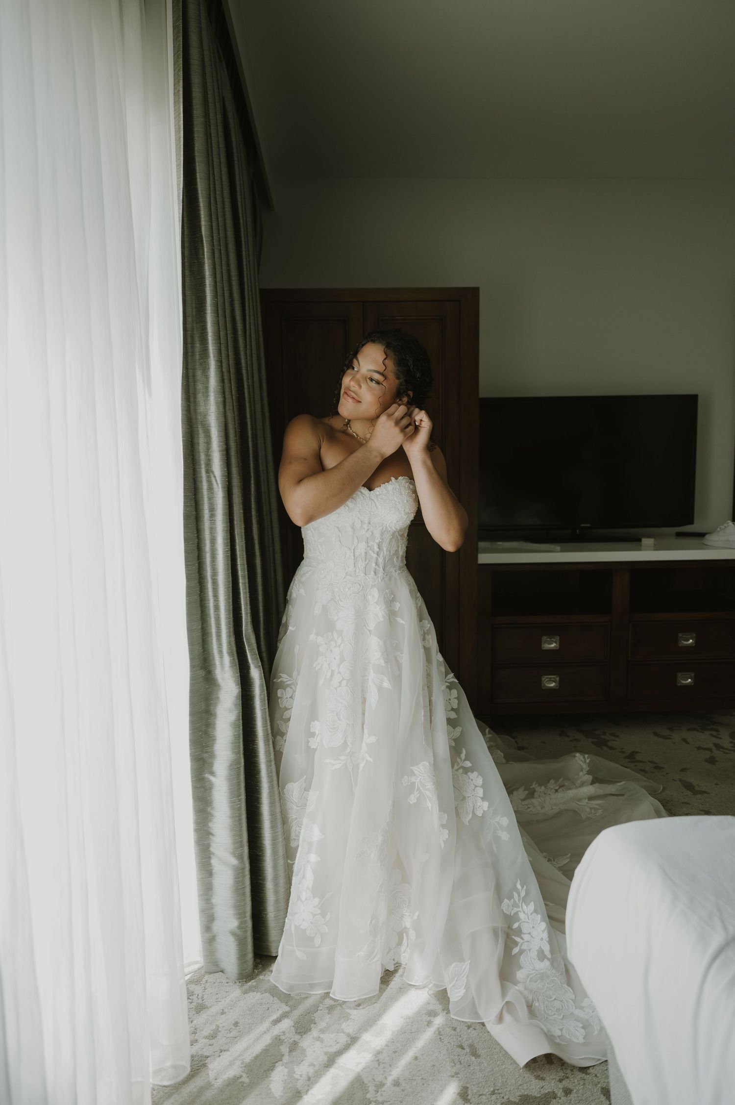 Bride in strapless lace A-line wedding gown adjusting earring by window in hotel room.