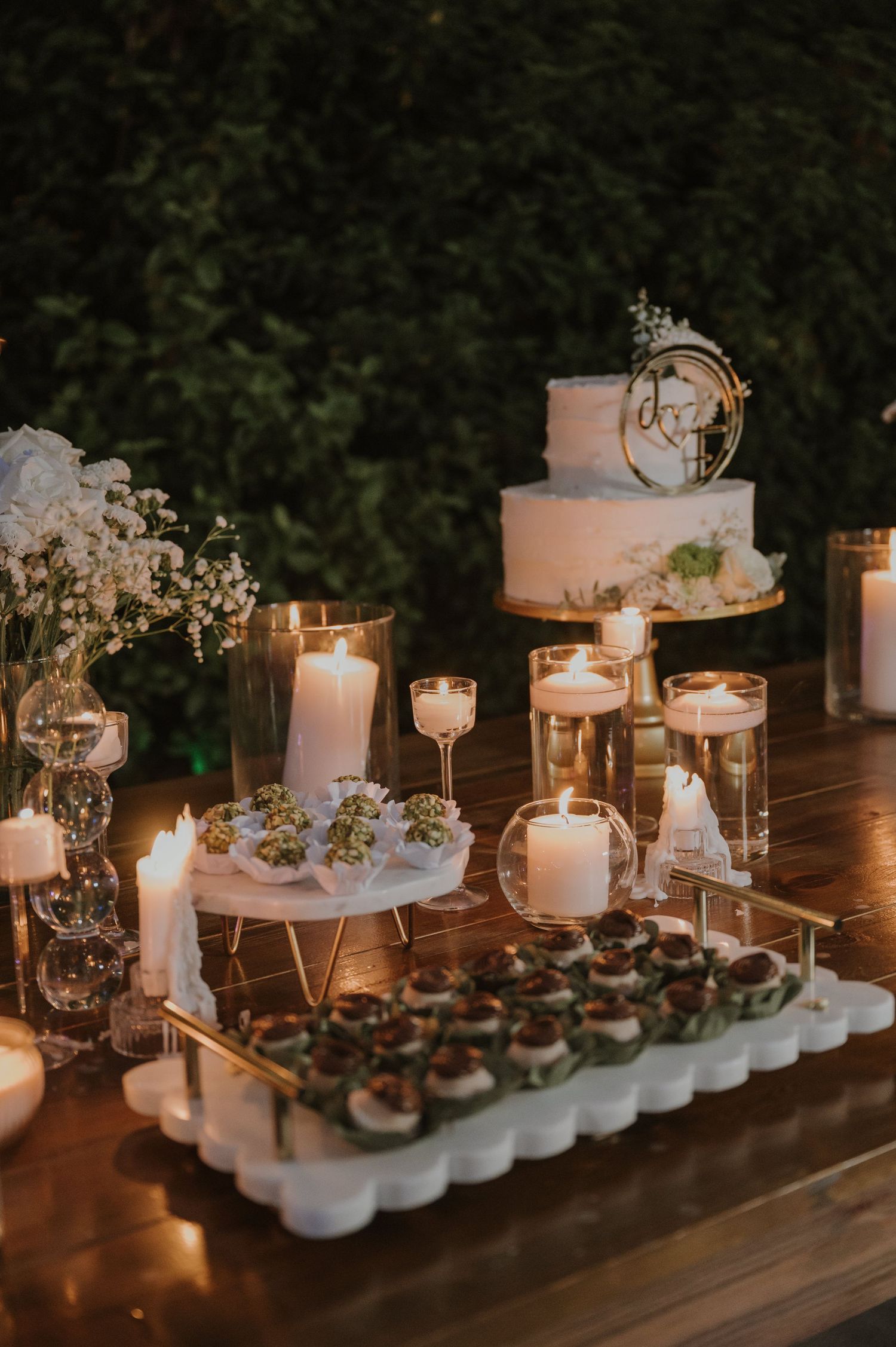Elegant wedding dessert table with white cake, candles, and treats against lush green foliage.