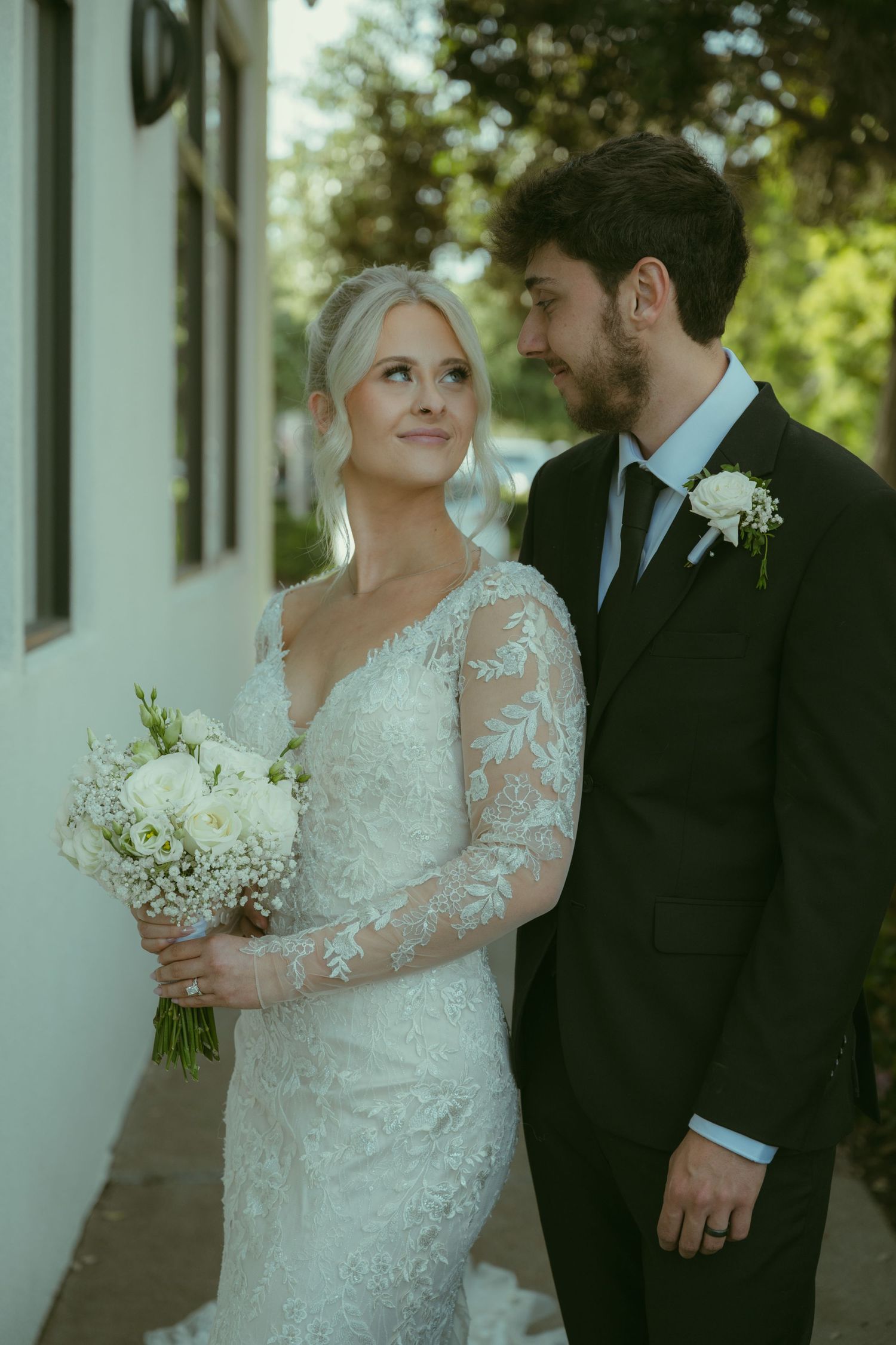 Bride in lace long-sleeve gown holds white bouquet, gazing at groom in black suit with boutonniere outdoors.