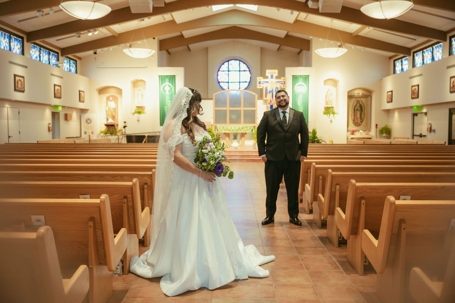 Bride in white gown and groom in black suit stand apart in church aisle, surrounded by wooden pews and green altar decor.