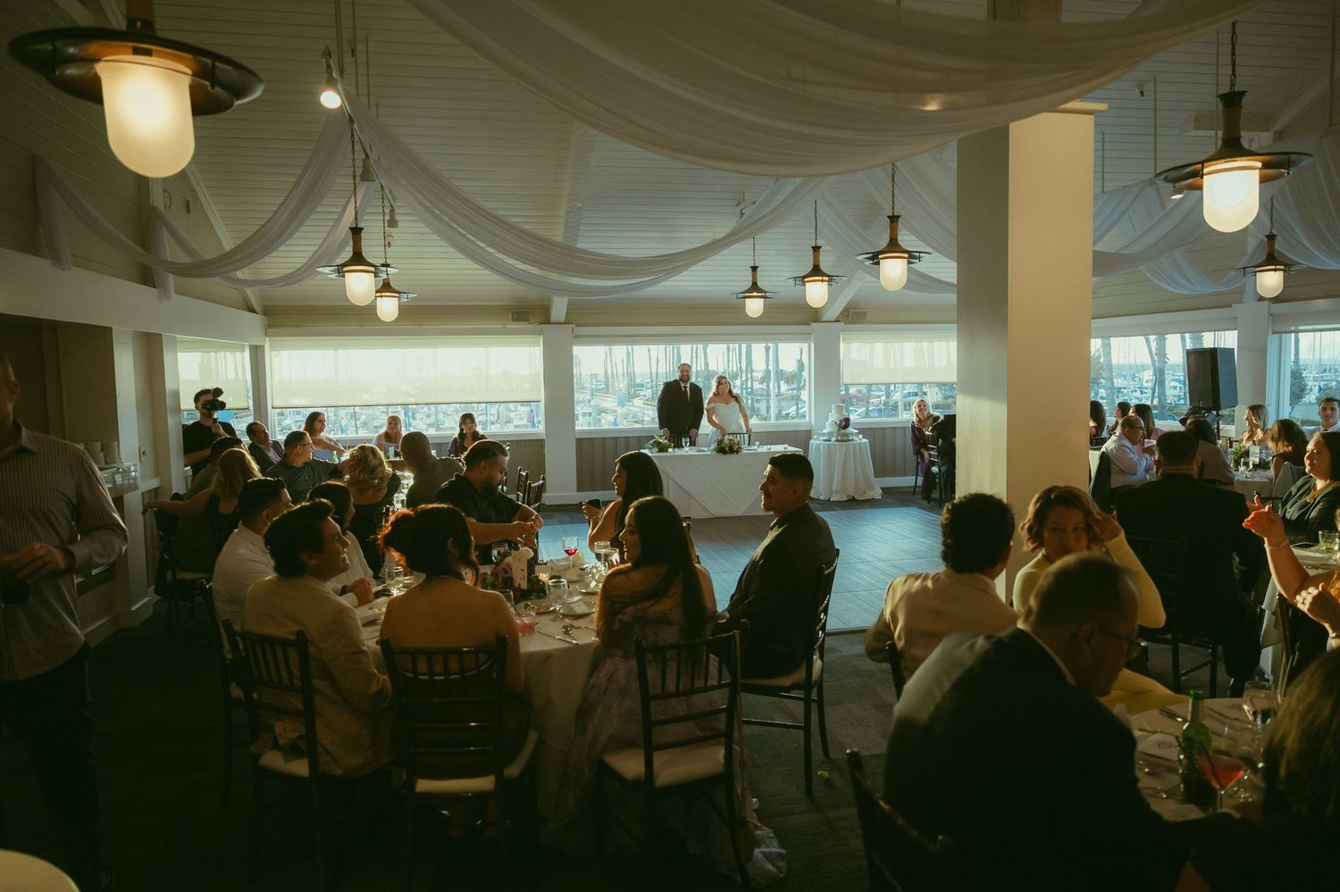 Wedding reception guests seated at round tables in elegant draped venue while couple stands at sweetheart table.