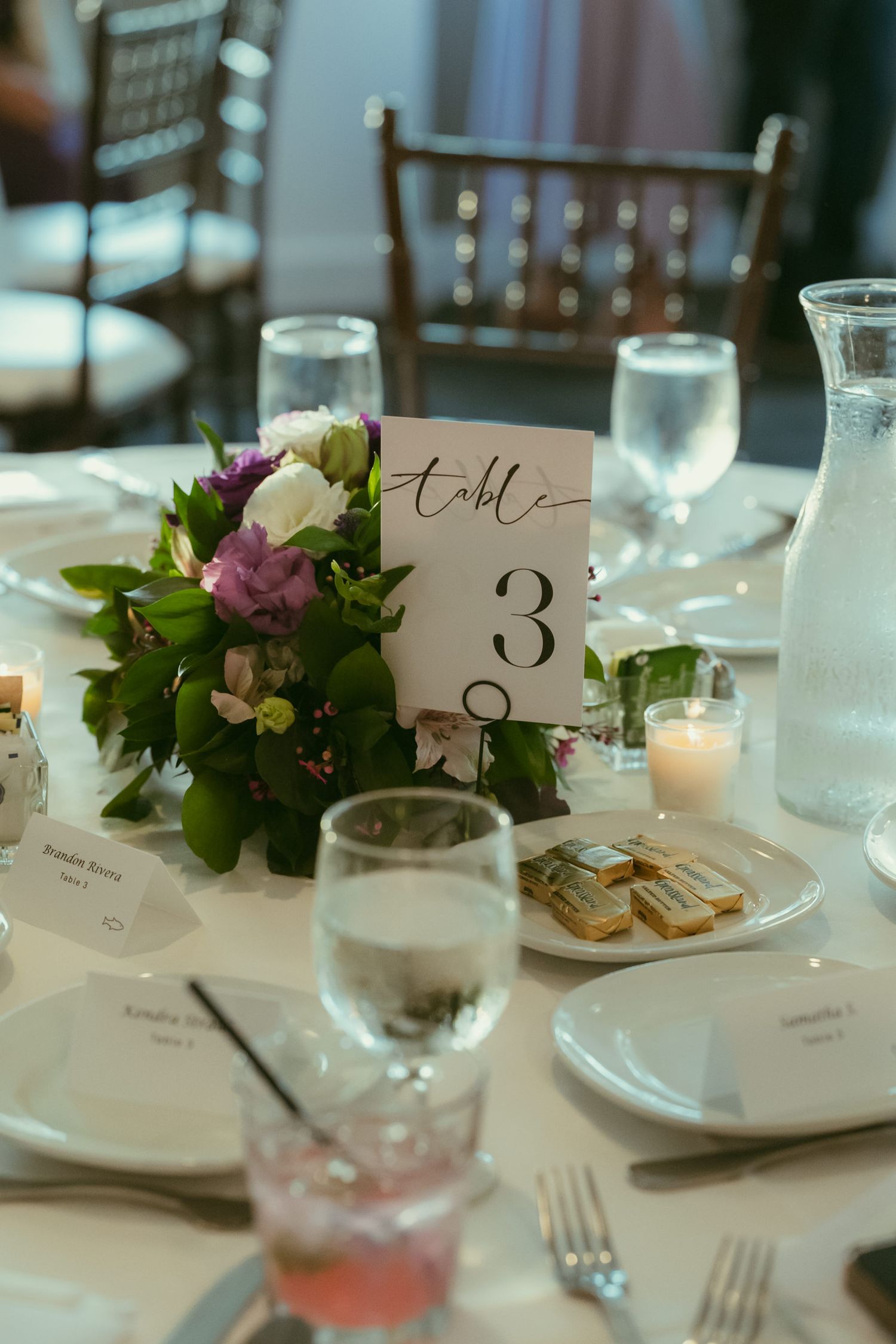 Elegant wedding reception table setting with floral centerpiece, table number 3 card, glassware, candles, and place settings.