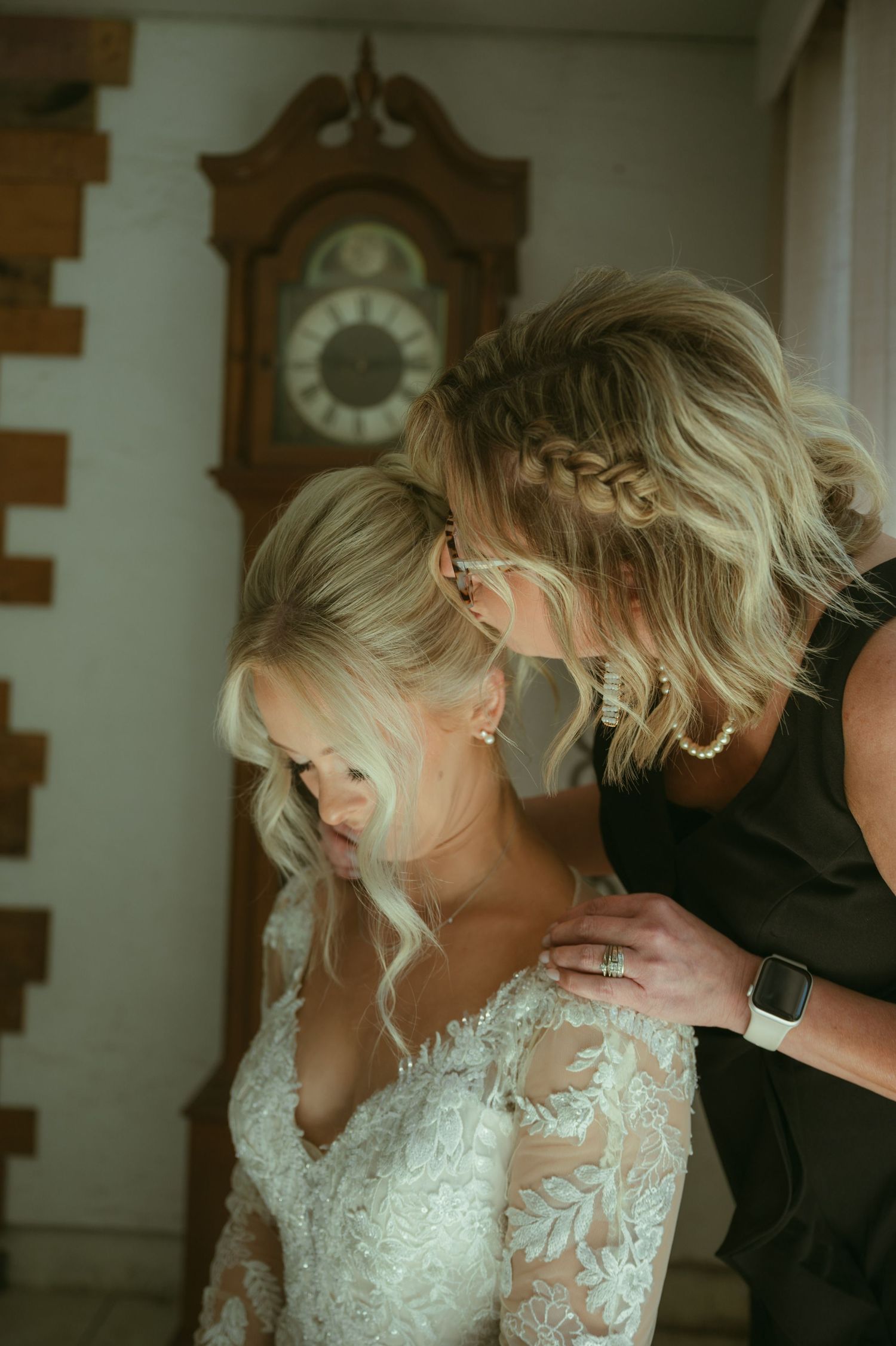 A woman helps a bride in a lace wedding gown prepare, with a vintage grandfather clock in the background.