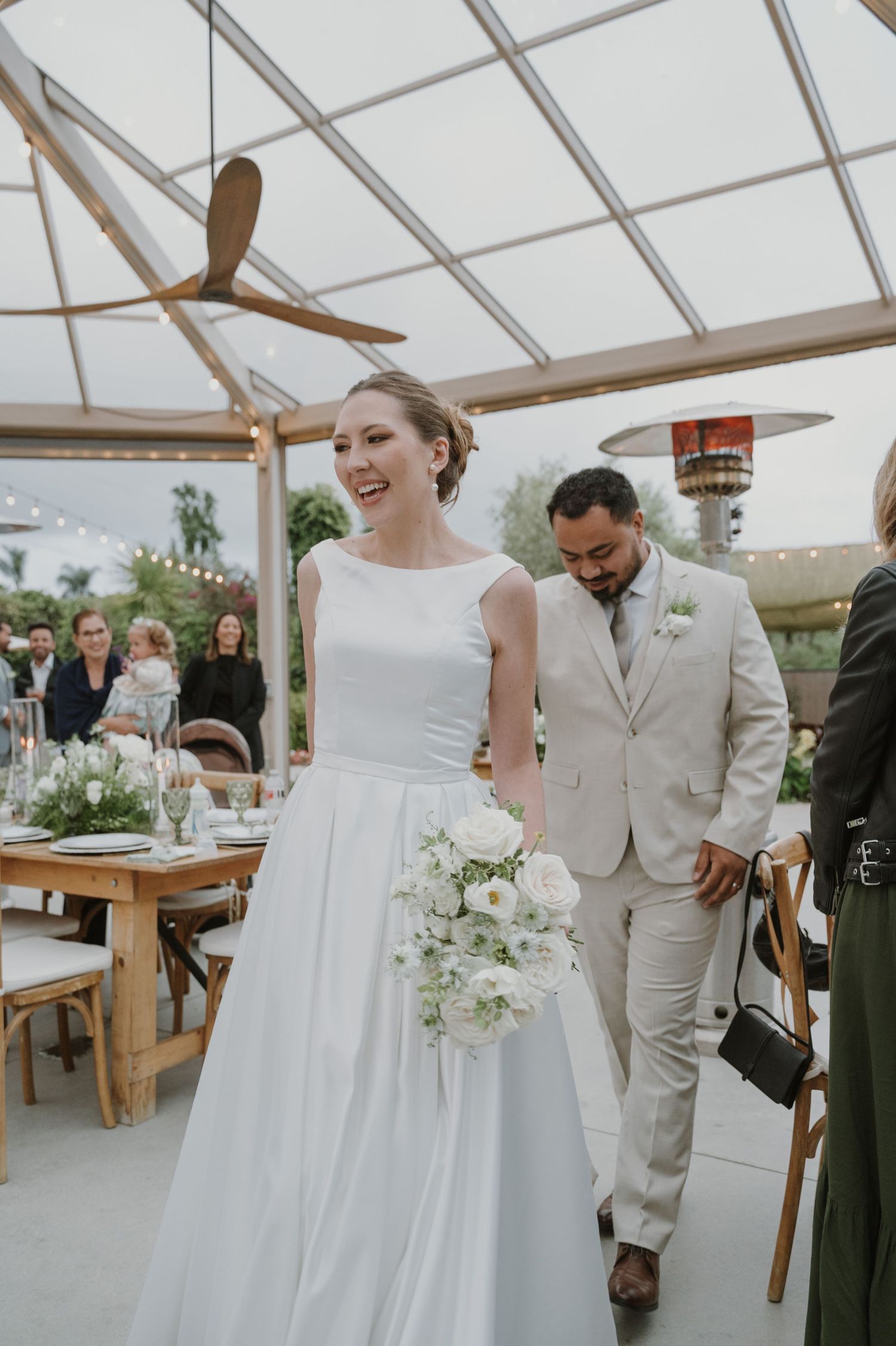 Smiling bride in white gown holding white bouquet walks with groom in beige suit at elegant wedding reception.