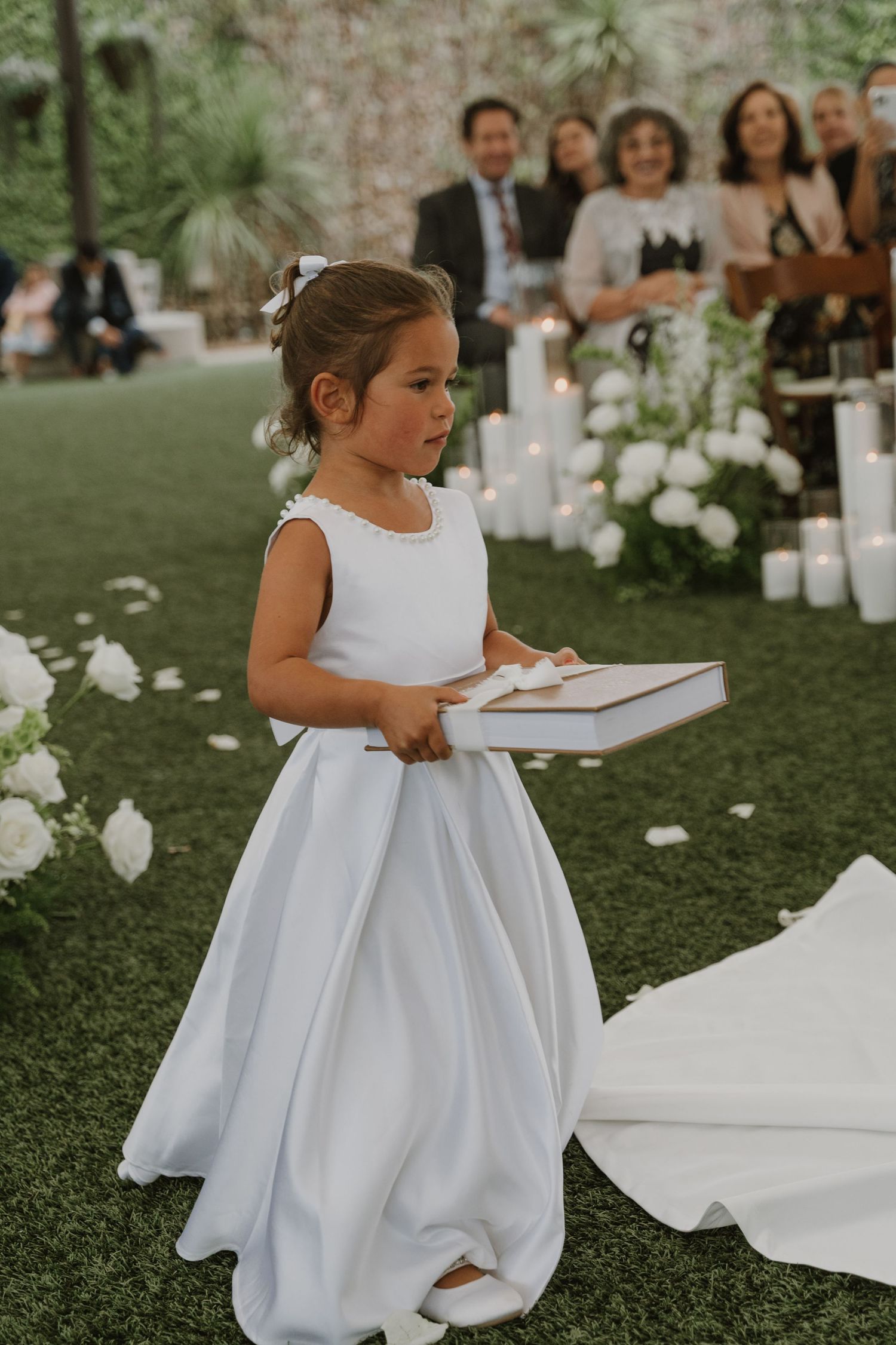 A young flower girl in a white dress carries a ring box down the aisle at an outdoor wedding ceremony.