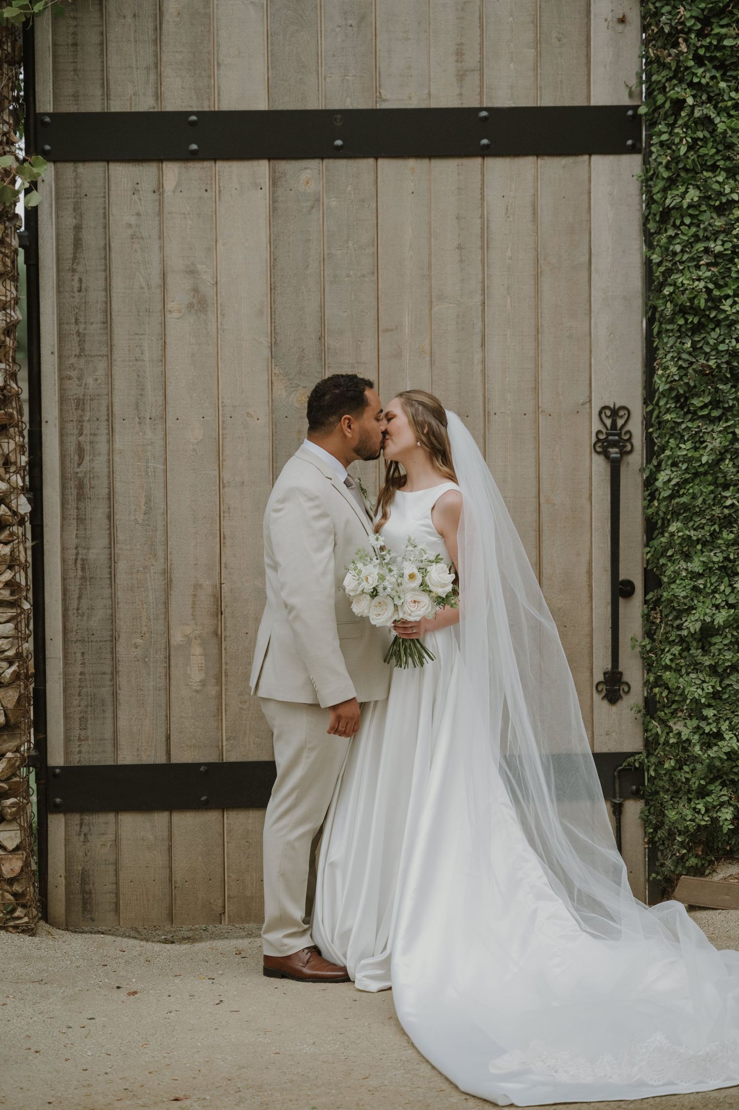 Bride and groom share a kiss in front of rustic wooden barn doors, bride holding white bouquet with long veil.