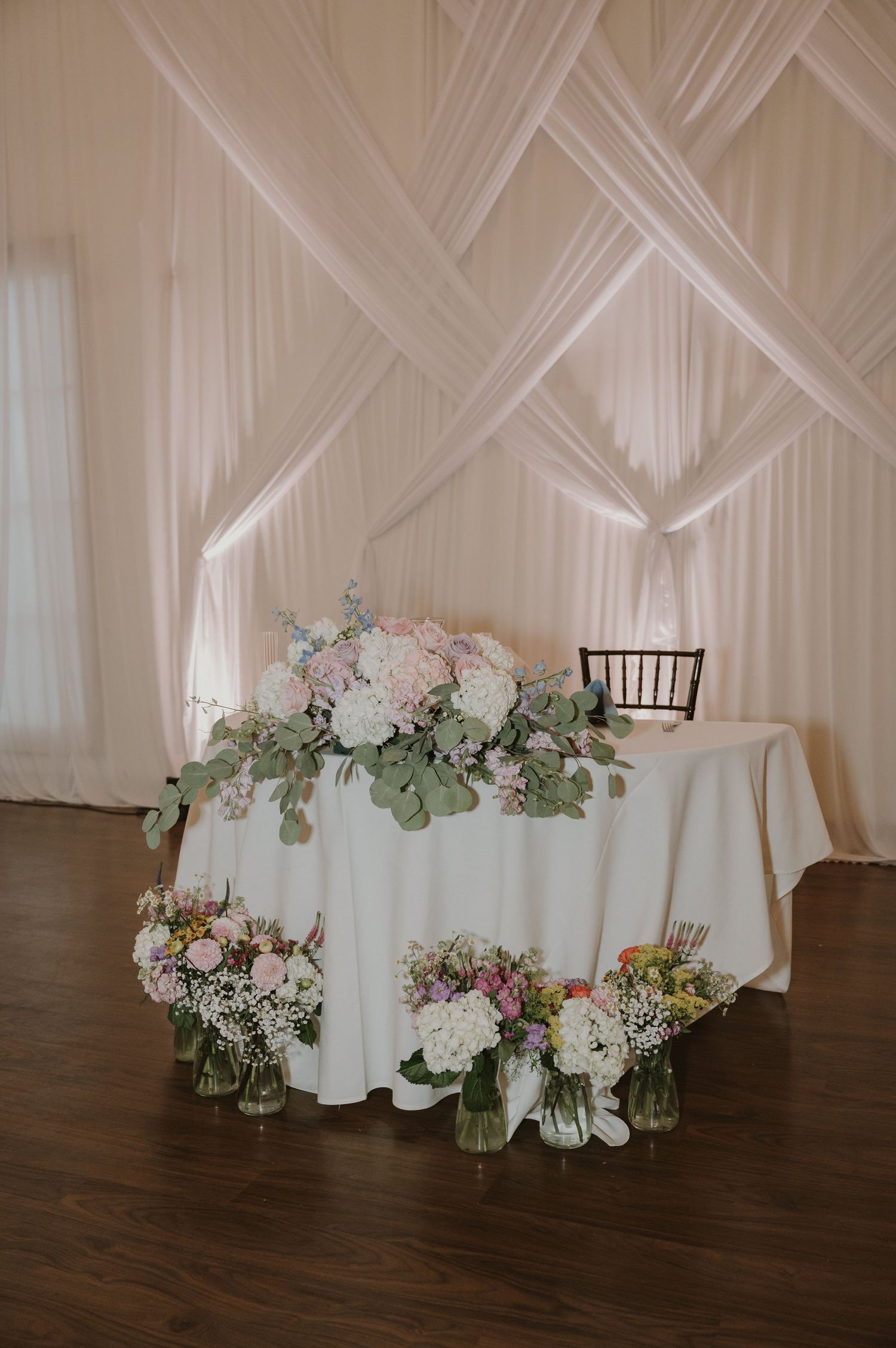 Wedding sweetheart table adorned with white hydrangeas and greenery against draped white backdrop.