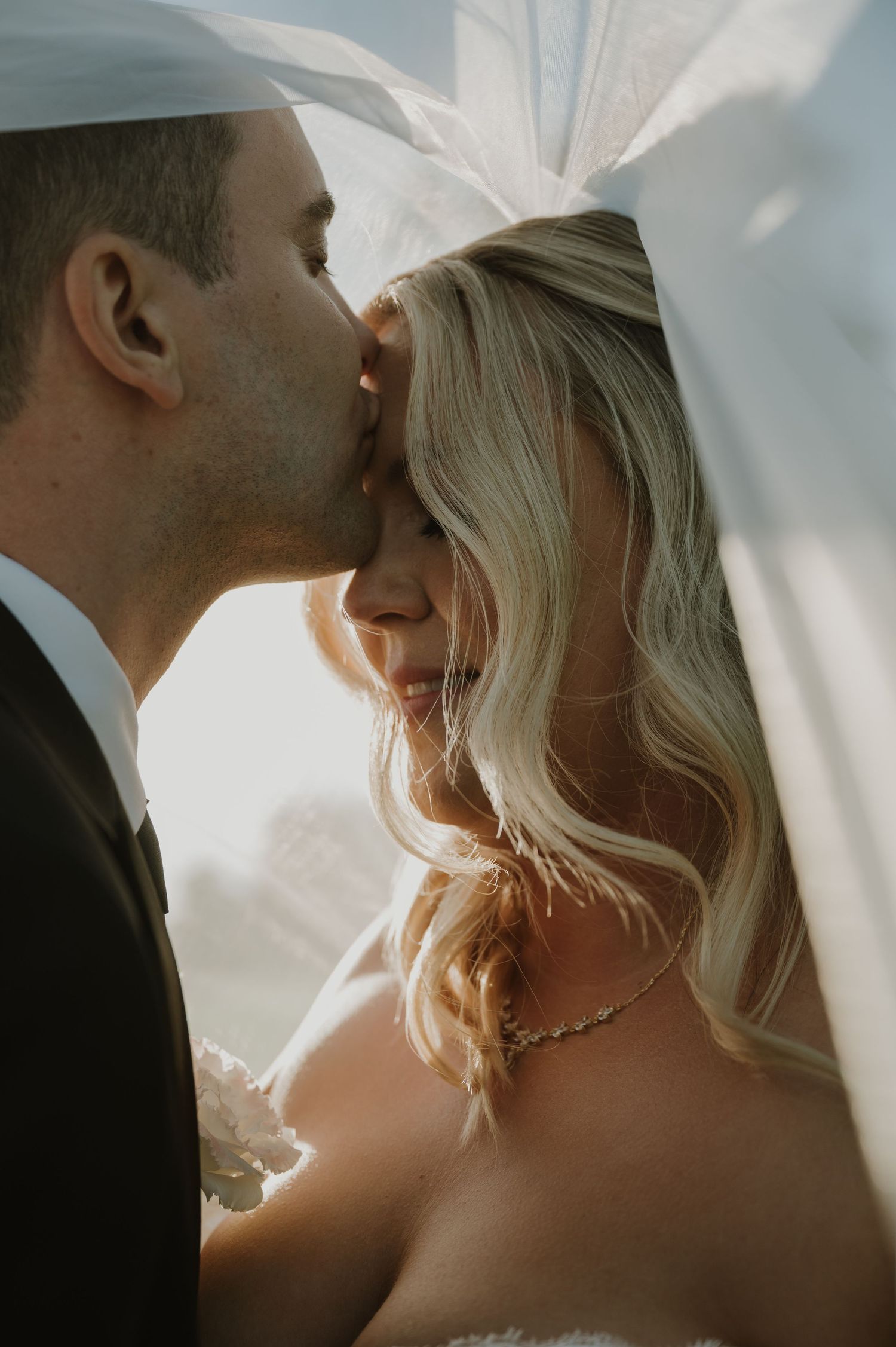 Groom kissing blonde bride on forehead under flowing veil in warm golden light.
