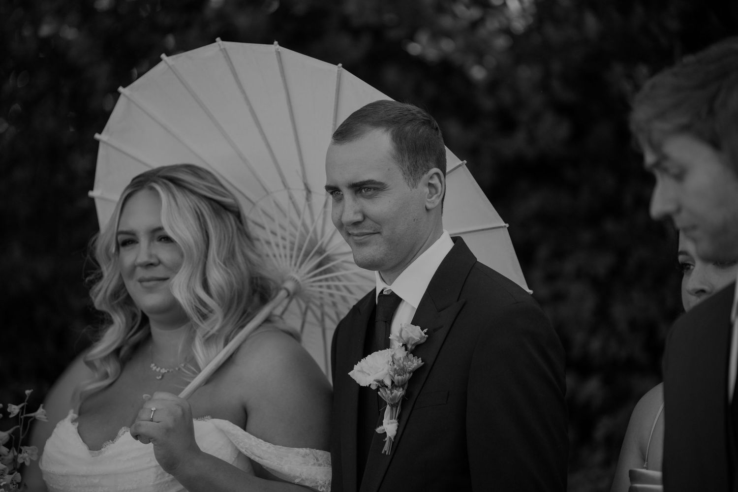 Black and white photo of smiling bride and groom holding white parasol during outdoor wedding ceremony.
