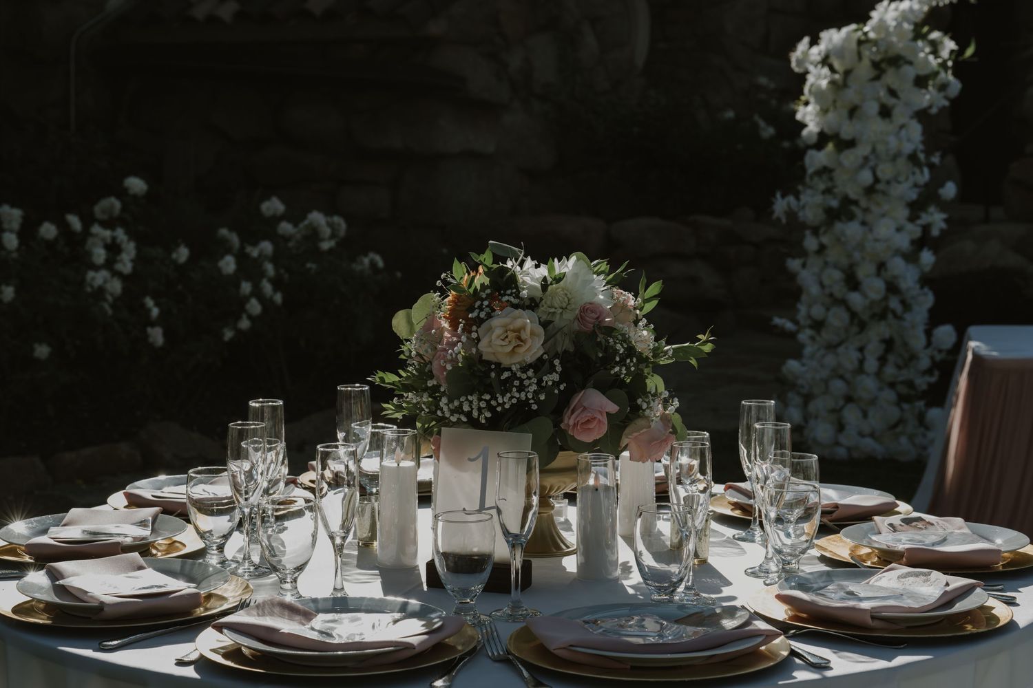 Romantic outdoor wedding reception table with floral centerpiece, gold charger plates, and crystal glassware.
