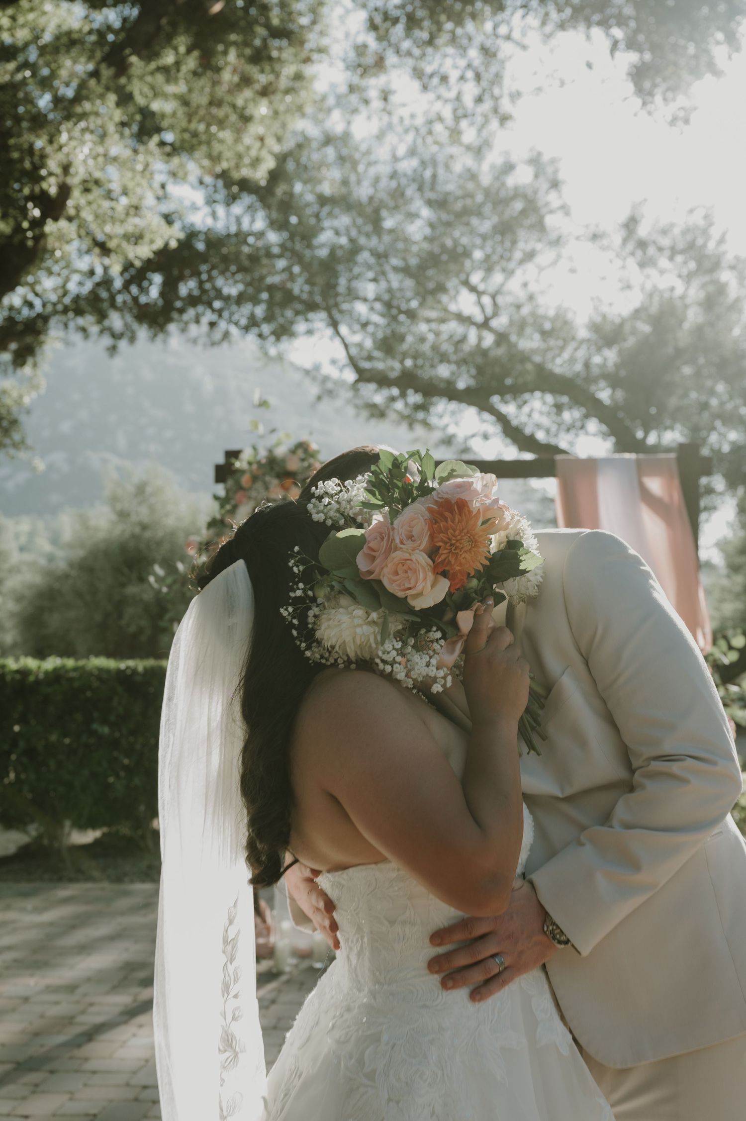 Bride and groom sharing a kiss, bouquet of pink roses visible, outdoor wedding ceremony with lush trees in background.