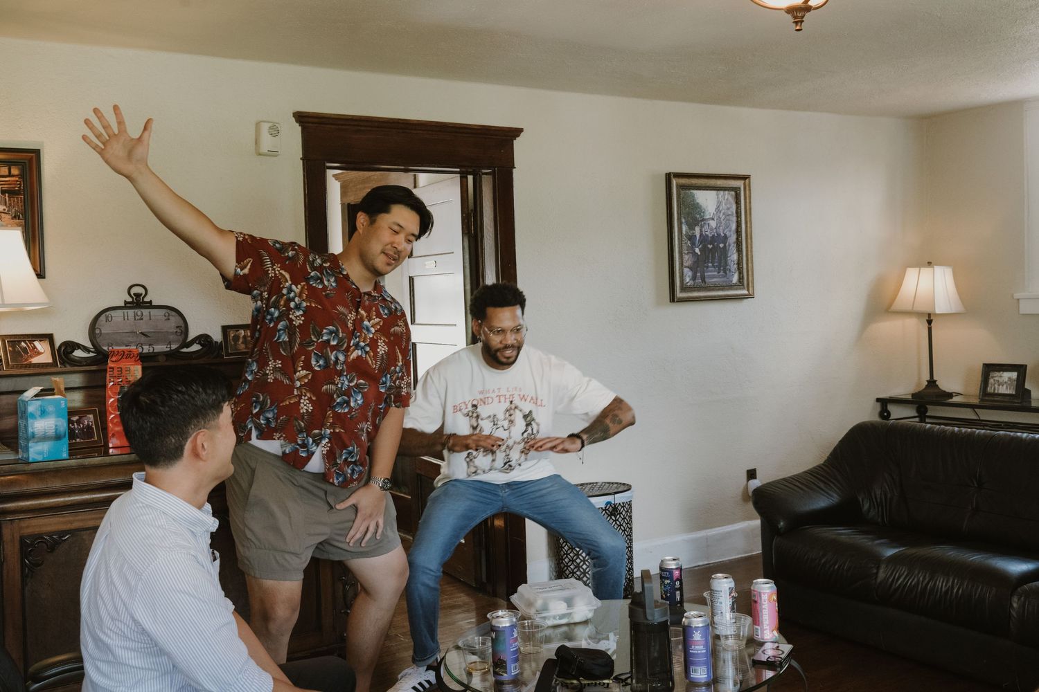 Three men celebrate in a living room, one dancing with raised arm in a floral shirt during a casual pre-wedding gathering.