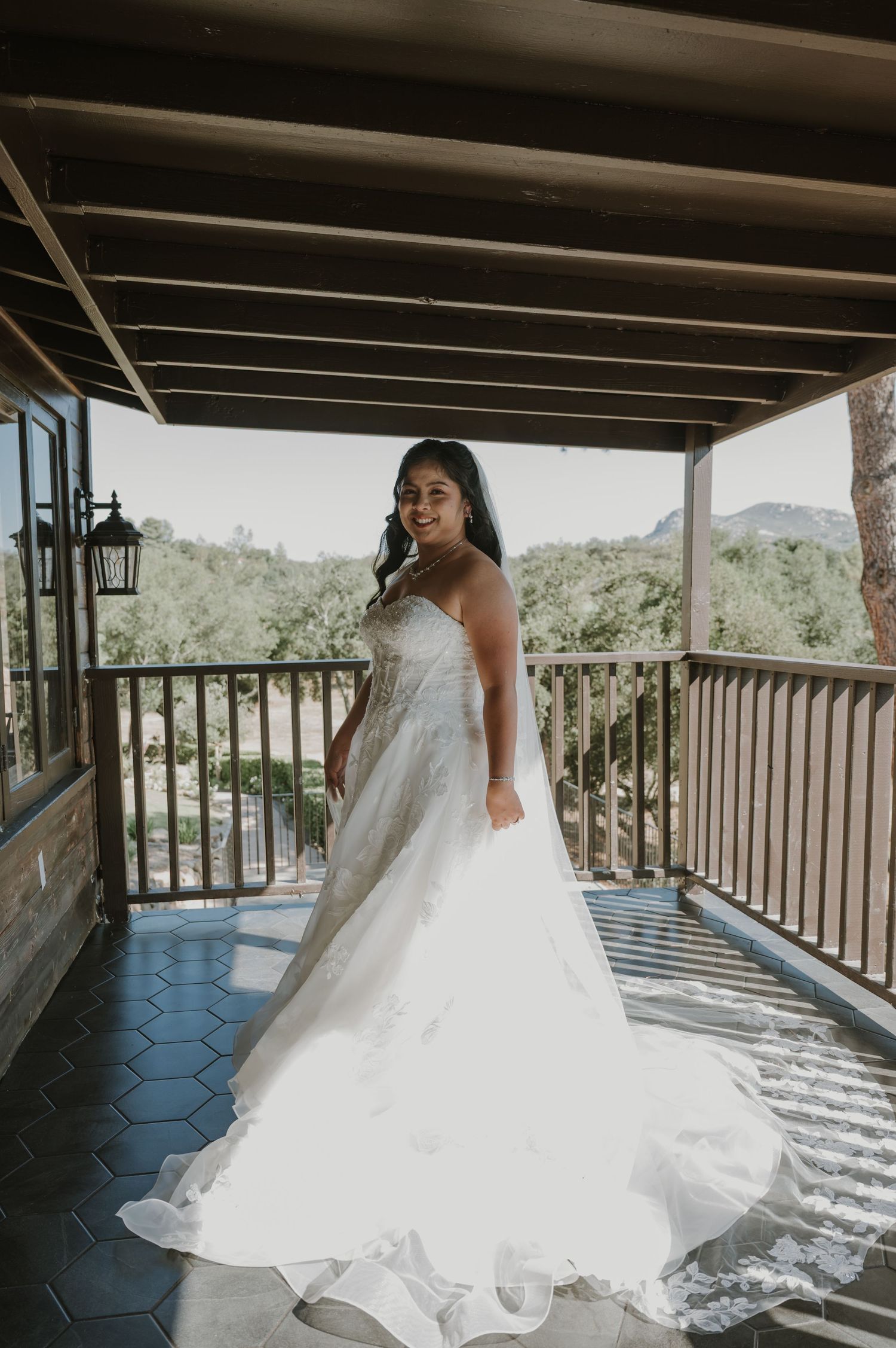 Bride in strapless ball gown wedding dress poses on rustic wooden balcony with scenic green hills behind her.