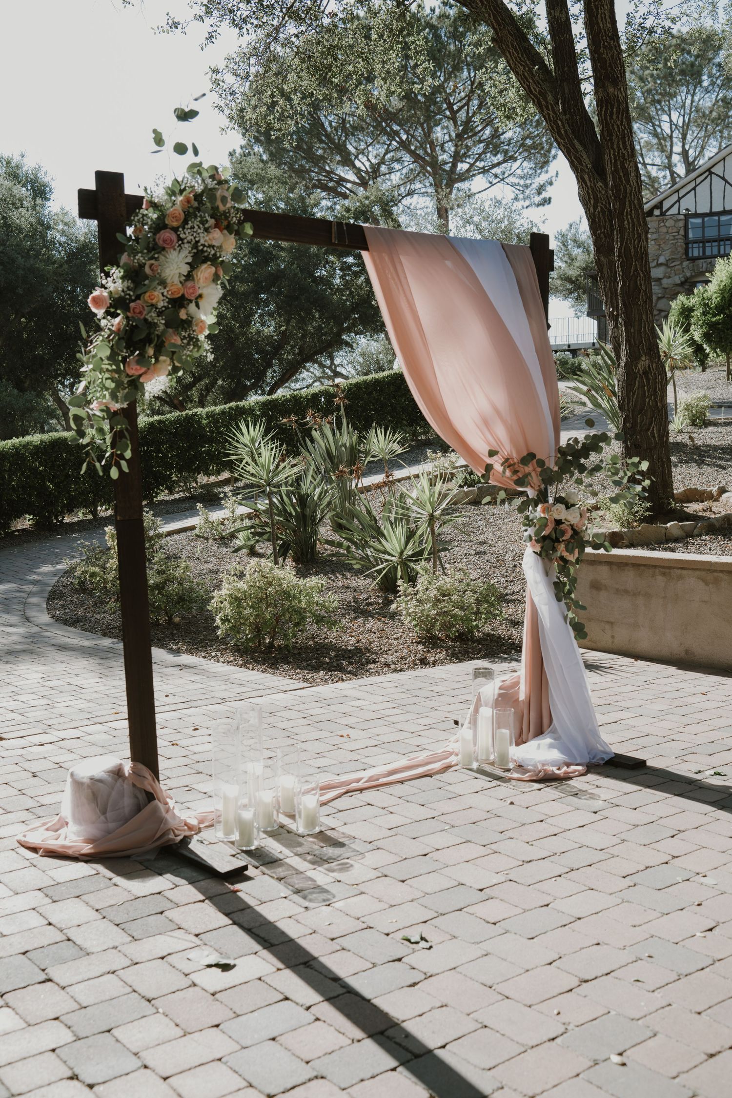 Rustic wooden wedding arch adorned with blush draping, pink florals, and candles on a brick patio.
