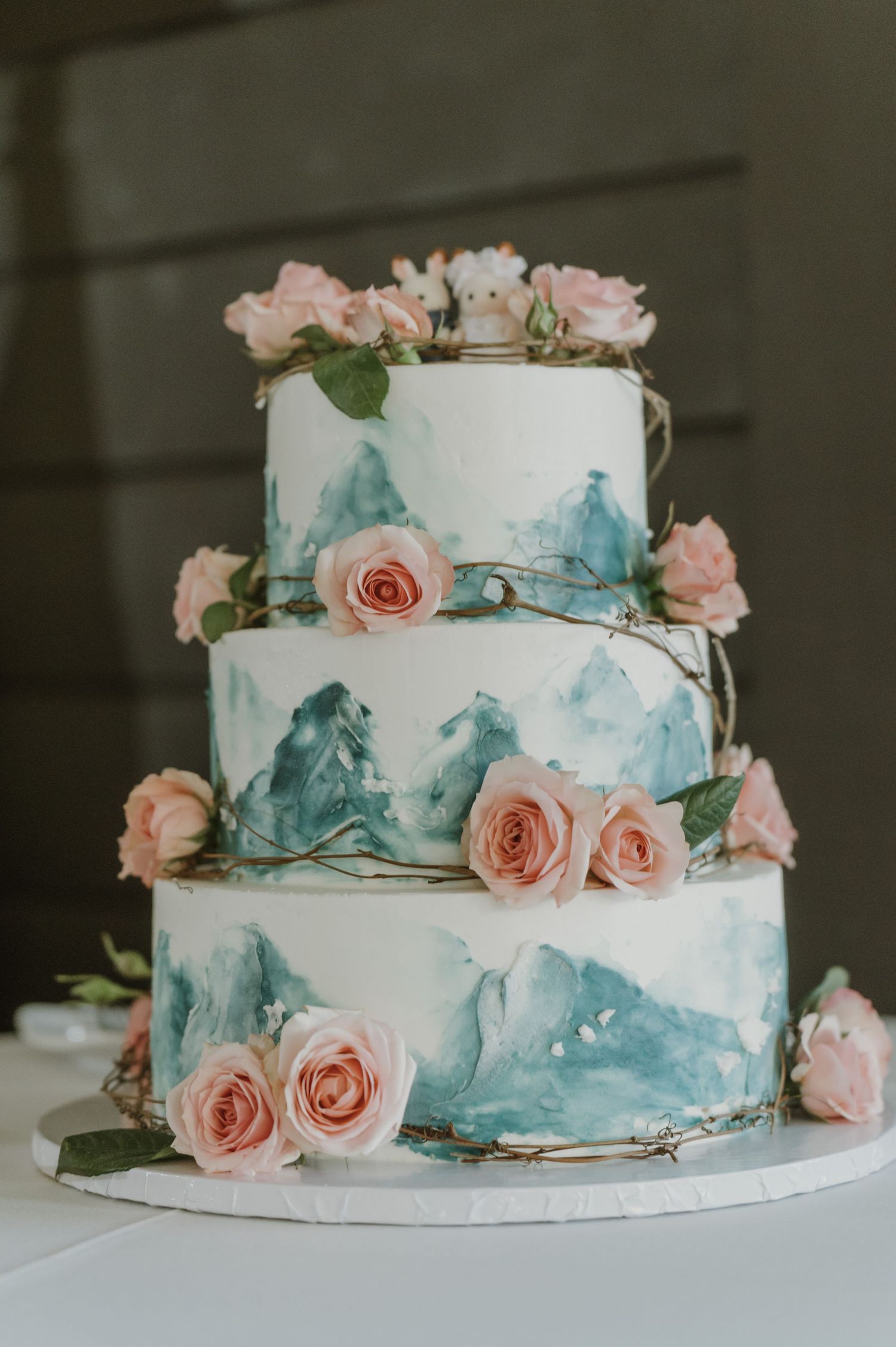 Three-tier white wedding cake with teal watercolor brushstrokes and fresh pink roses on each layer.