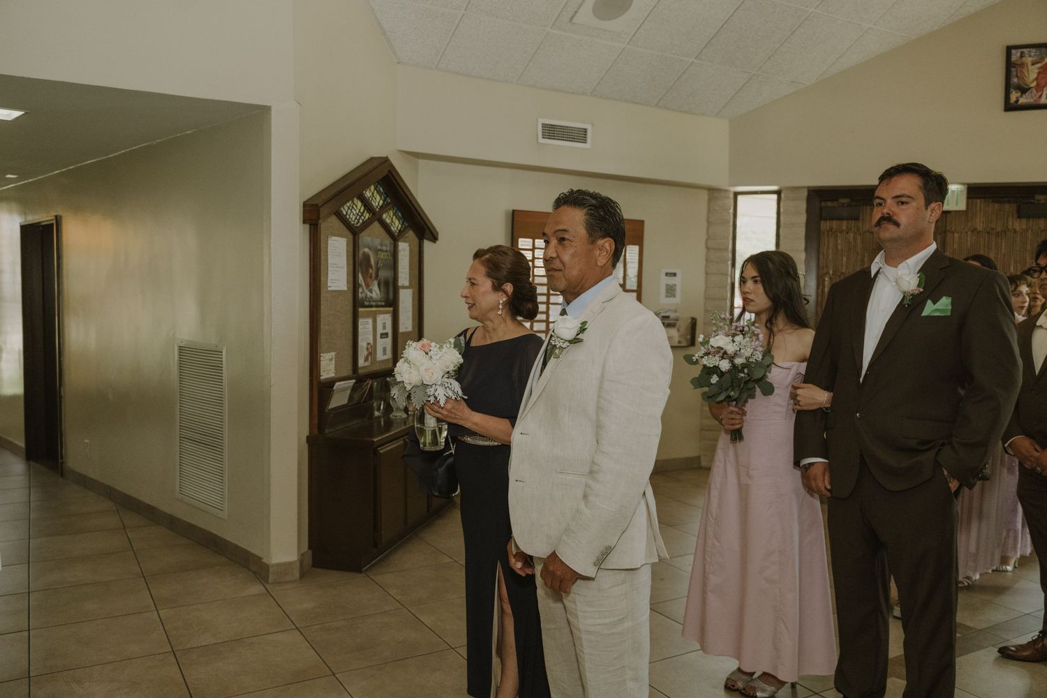 Wedding party members holding bouquets gather in a church hallway before the ceremony.