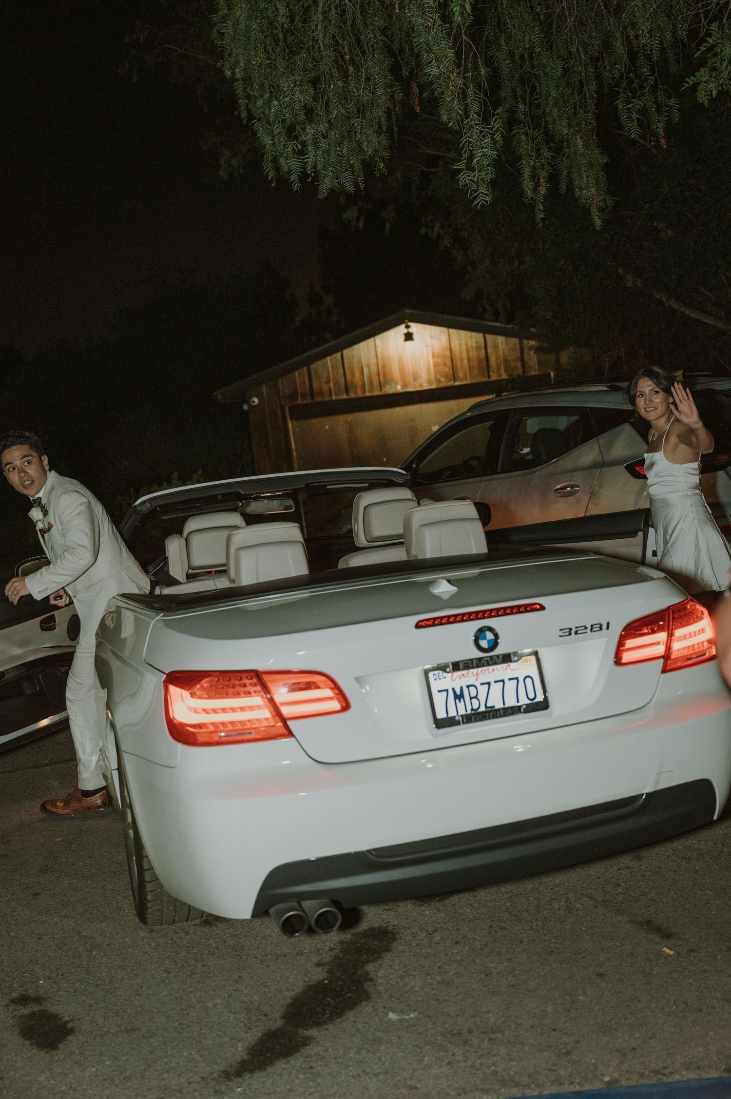Bride and groom pose beside white BMW 328i convertible during nighttime wedding getaway.